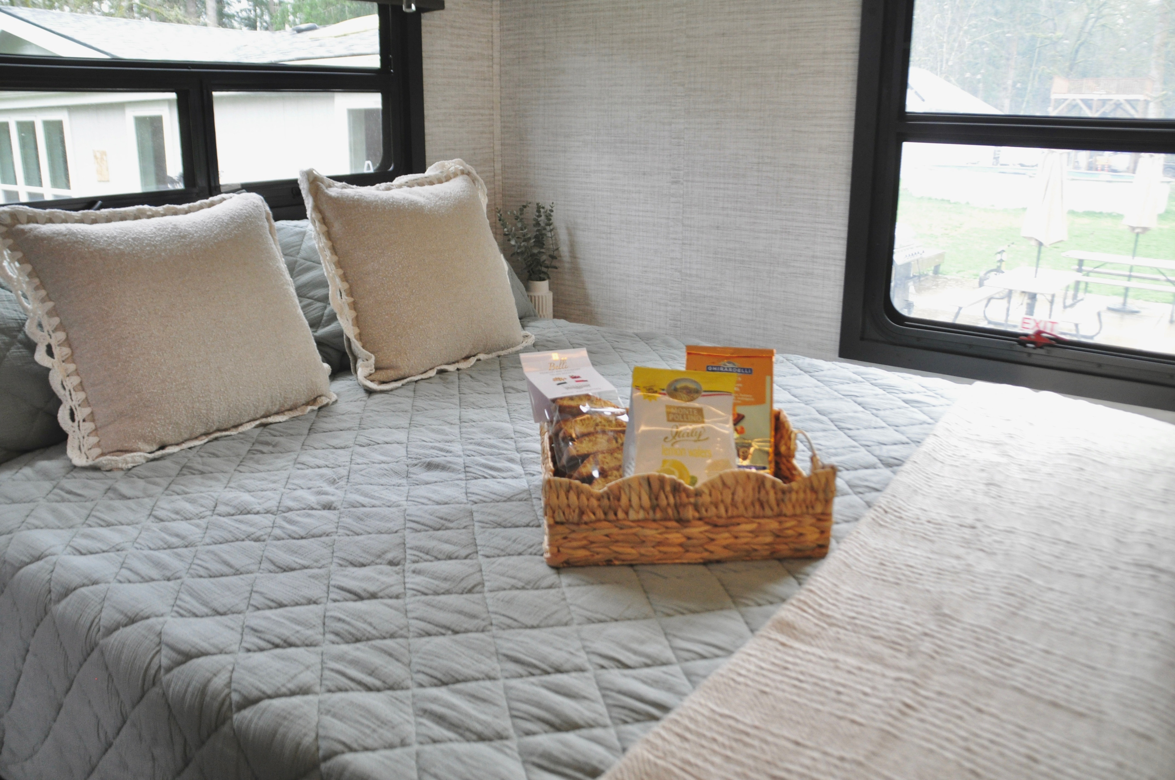 Cozy RV rental bed with light bedding, decorative pillows, and a welcome snack basket, offering a view of the outdoors.. Jayco Greyhawk 31F Bunkhouse 2025