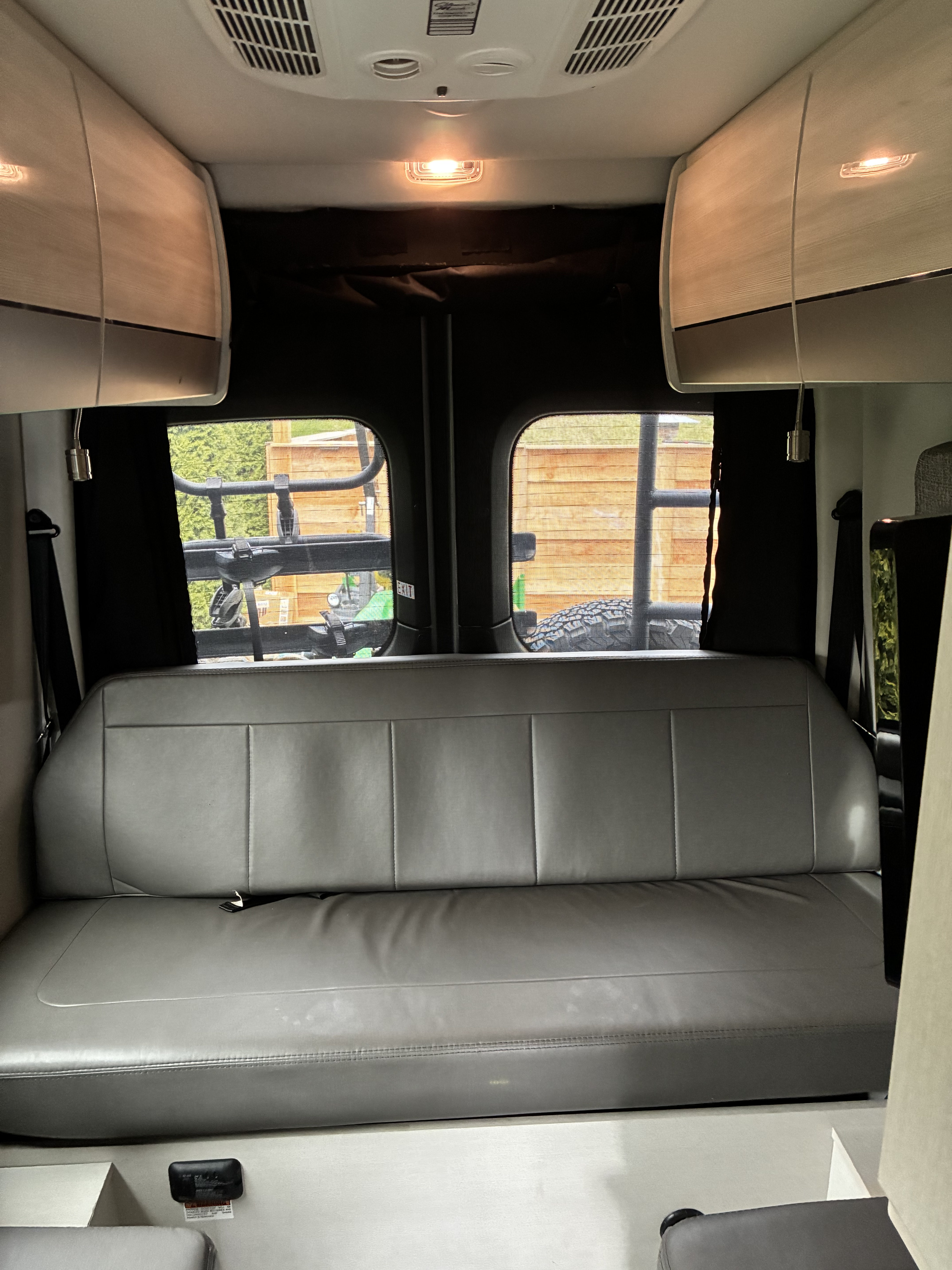 RV rental interior: A spacious grey bench seat and rear windows with outdoor views under clean overhead storage cabinets.. Thor Sanctuary 2022