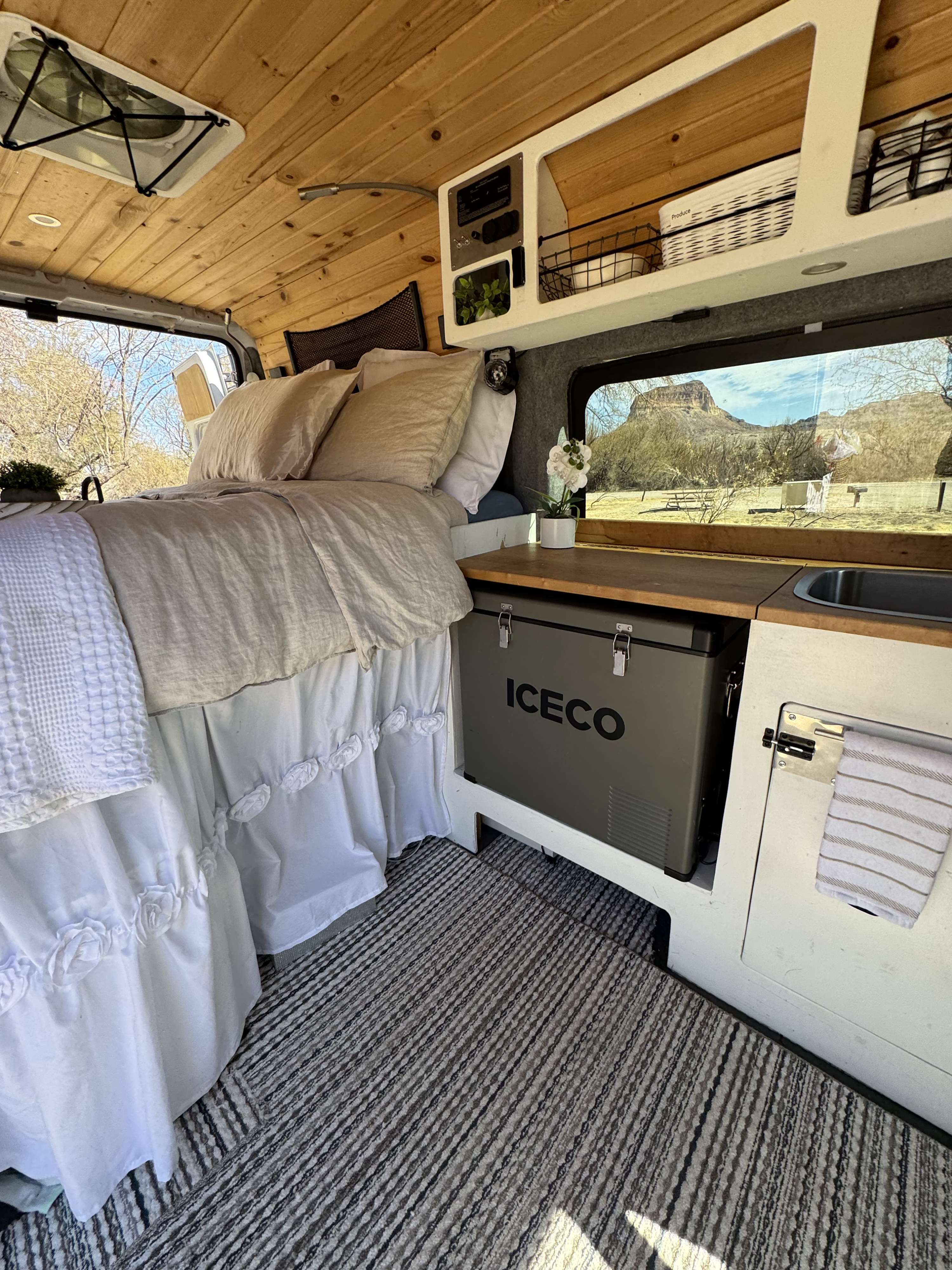 Cozy camper van RV rental interior: plush bed, Iceco fridge, sink, wood ceiling, and a scenic window view.. Dodge Ram 1500 2019