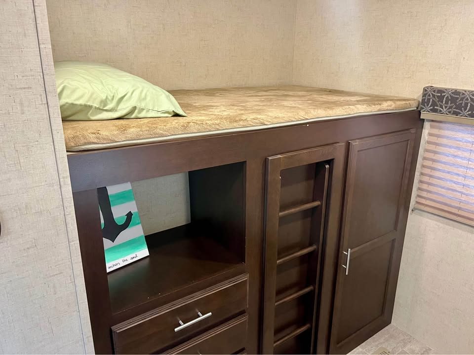 This RV rental bunk features a comfortable bed with a light green pillow, integrated dark wood storage, and a built-in ladder.. KZ Sportsman 2018