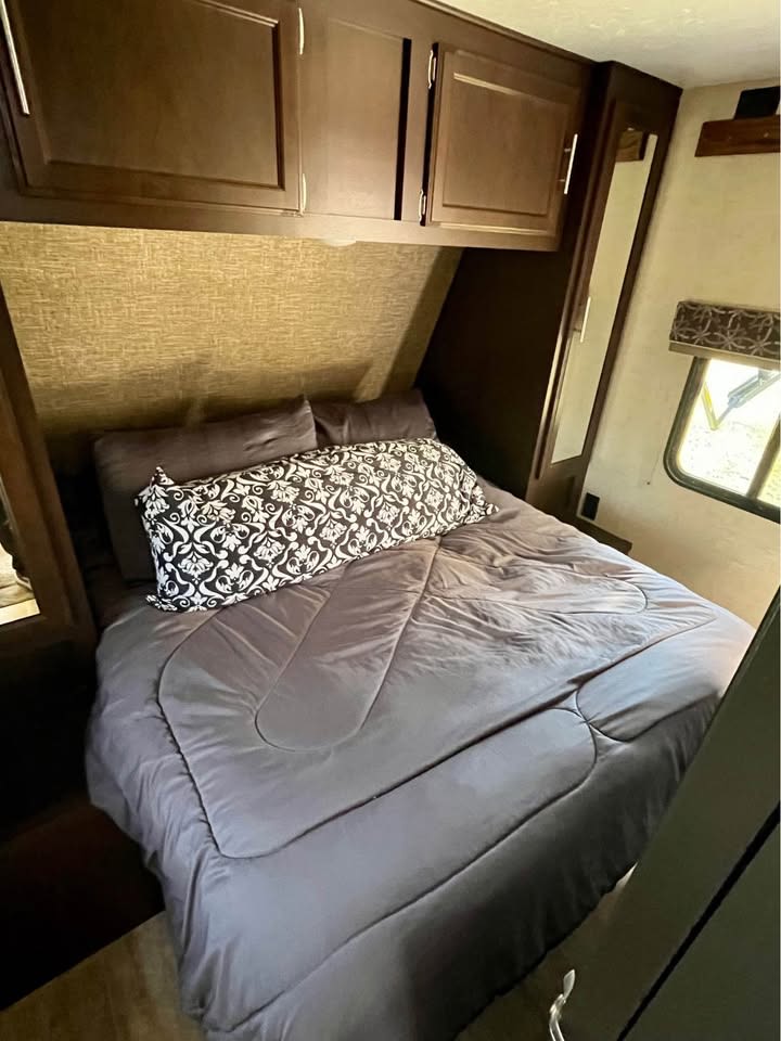 An RV rental bedroom with a comfortable bed, gray bedding, decorative pillow, and overhead wood cabinets.. KZ Sportsman 2018