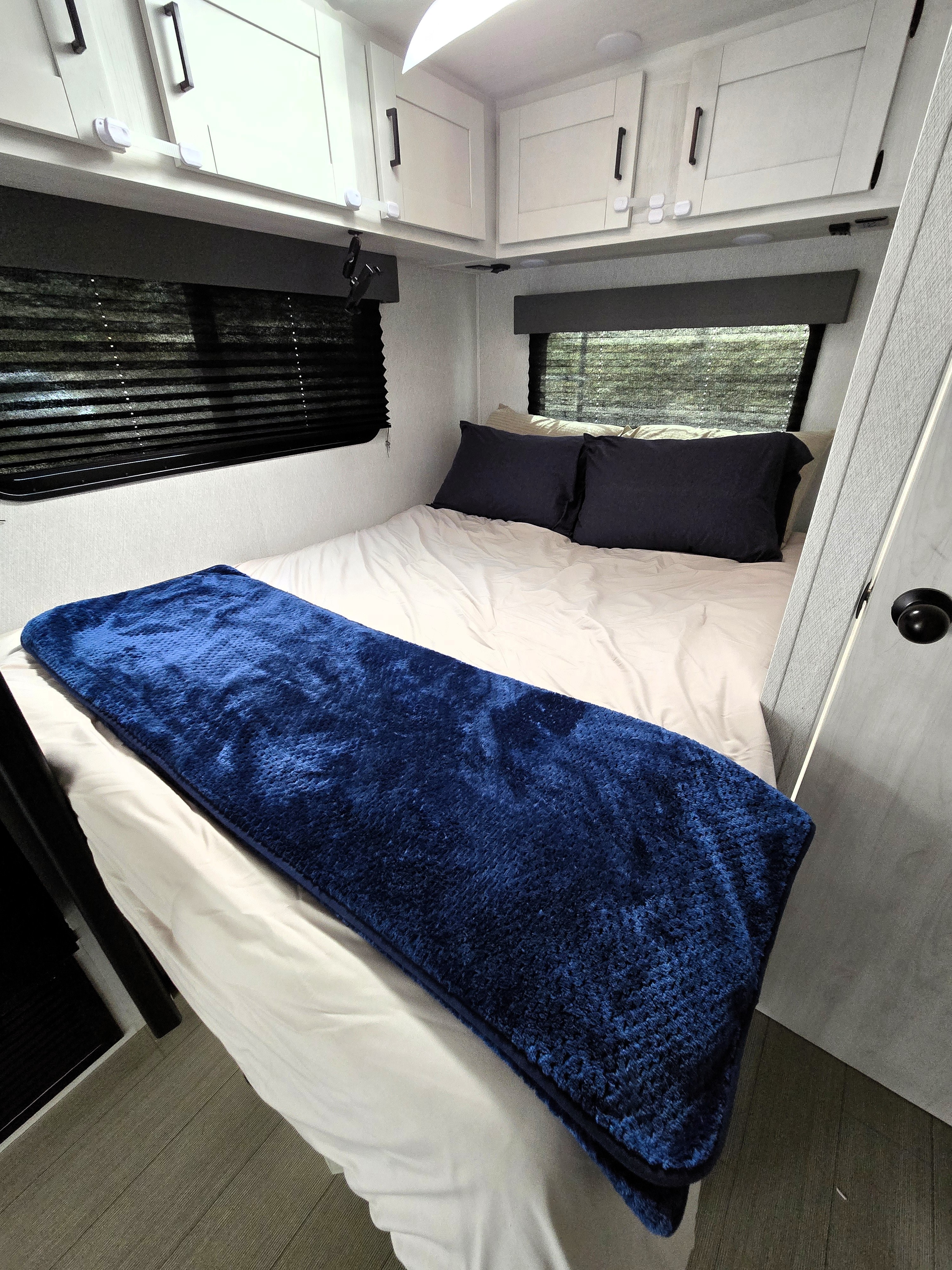 Cozy RV rental bedroom with blue blanket, white sheets, and ample storage in overhead cabinets.. East to west Entrada 2025