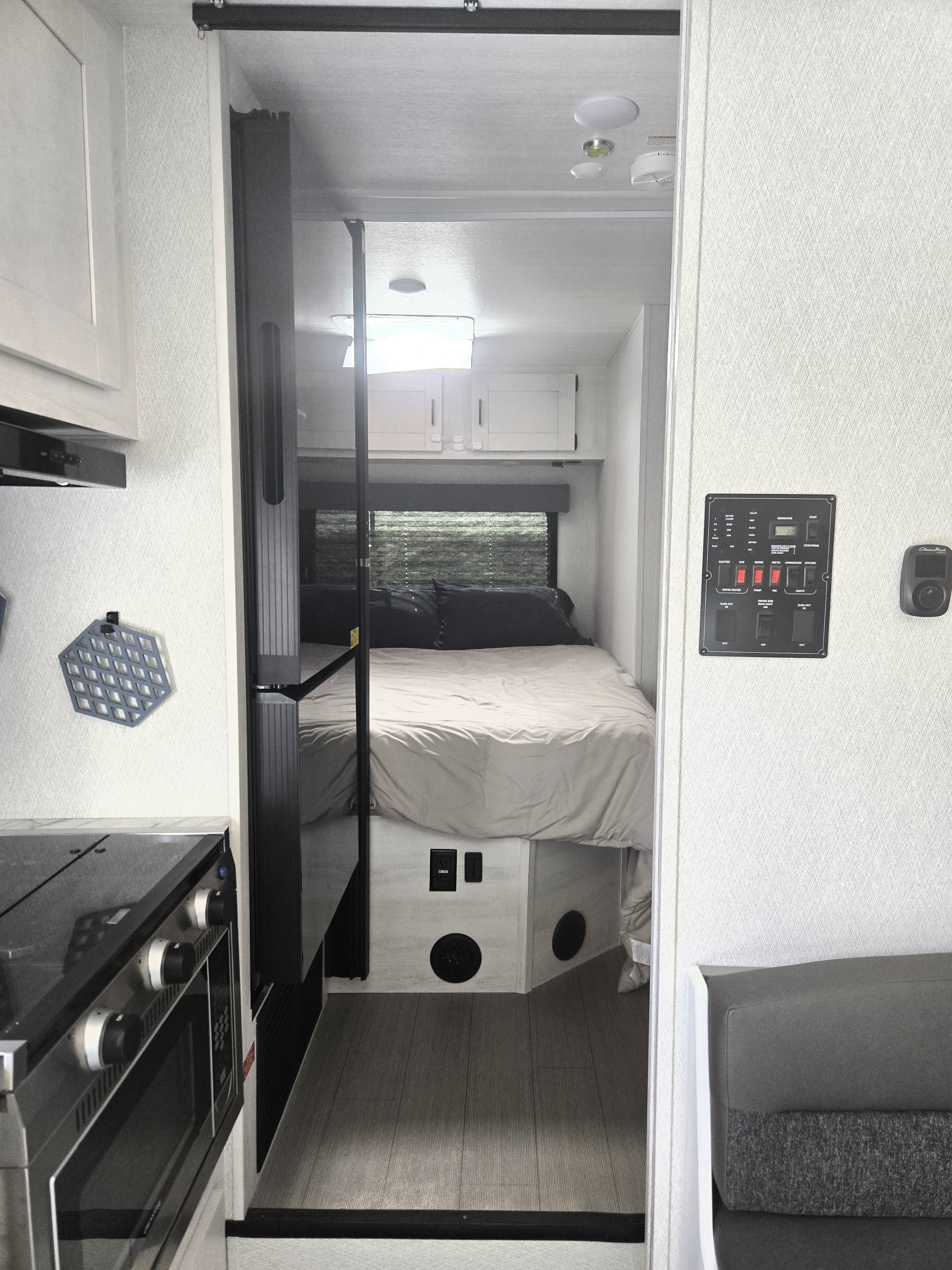 This rv rental interior features a kitchen counter, oven, and control panel, leading to a bedroom with a neatly made bed and window.. East to west Entrada 2025