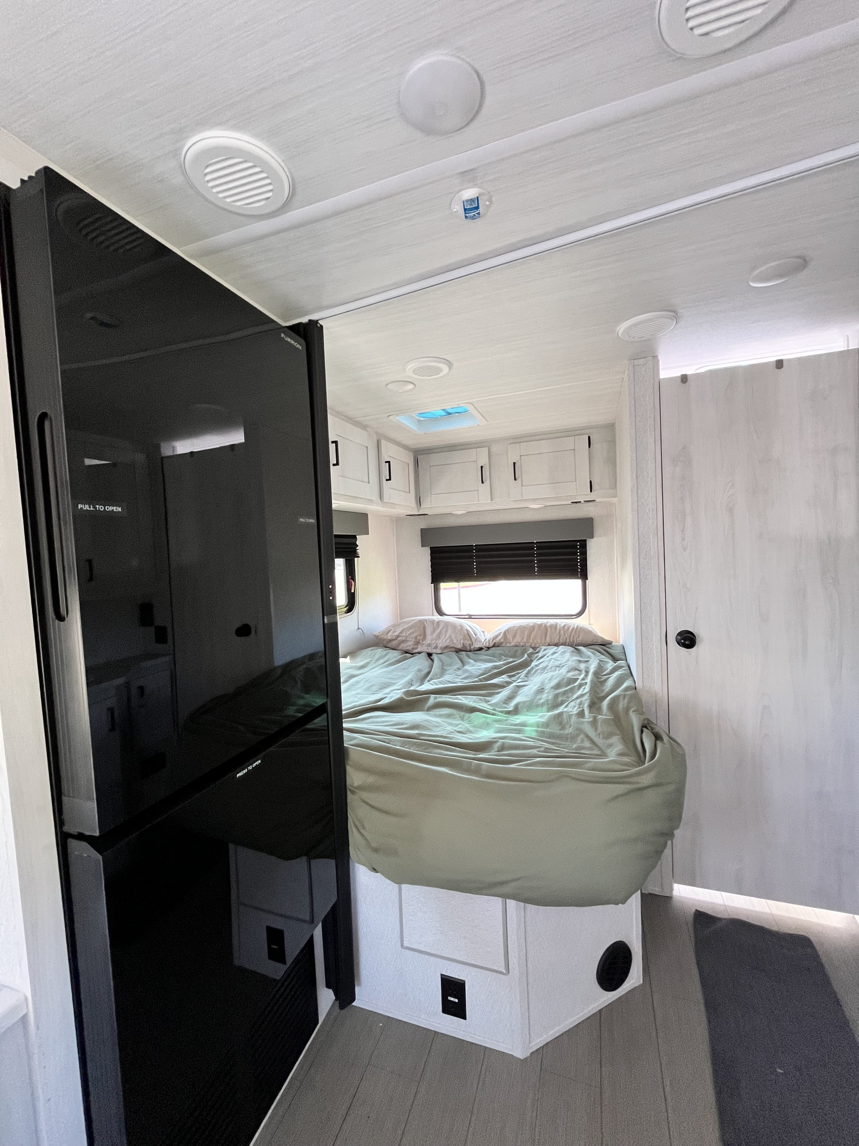 This RV rental's bright bedroom shows a comfortable bed, overhead cabinets, and a tall dark refrigerator unit.. East to West Entrada 210NS 2025