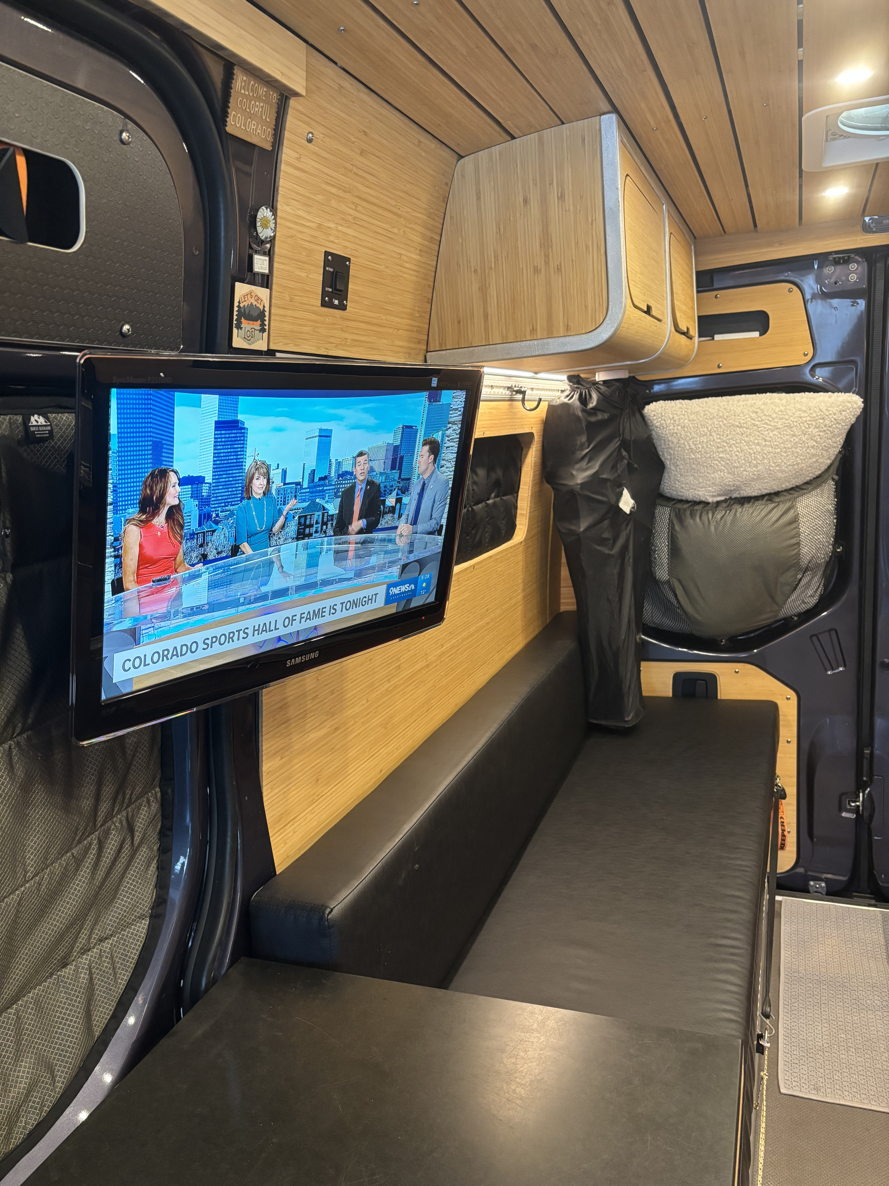 Cozy RV rental interior with bamboo walls, a TV displaying news, and comfortable black seating, ideal for road trips.. Mercedes-Benz Sprinter RV Motorhome Campervan 2023