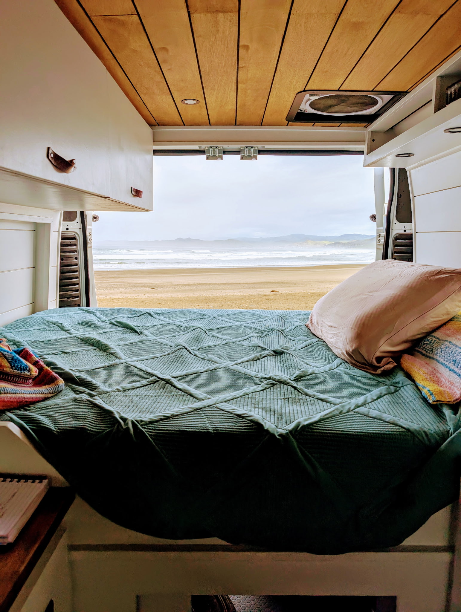 View from an RV rental's bed looking out to a sandy beach and ocean waves. Cozy van interior, wooden ceiling, and coastal scenery.. Dodge Ram 1500 2014
