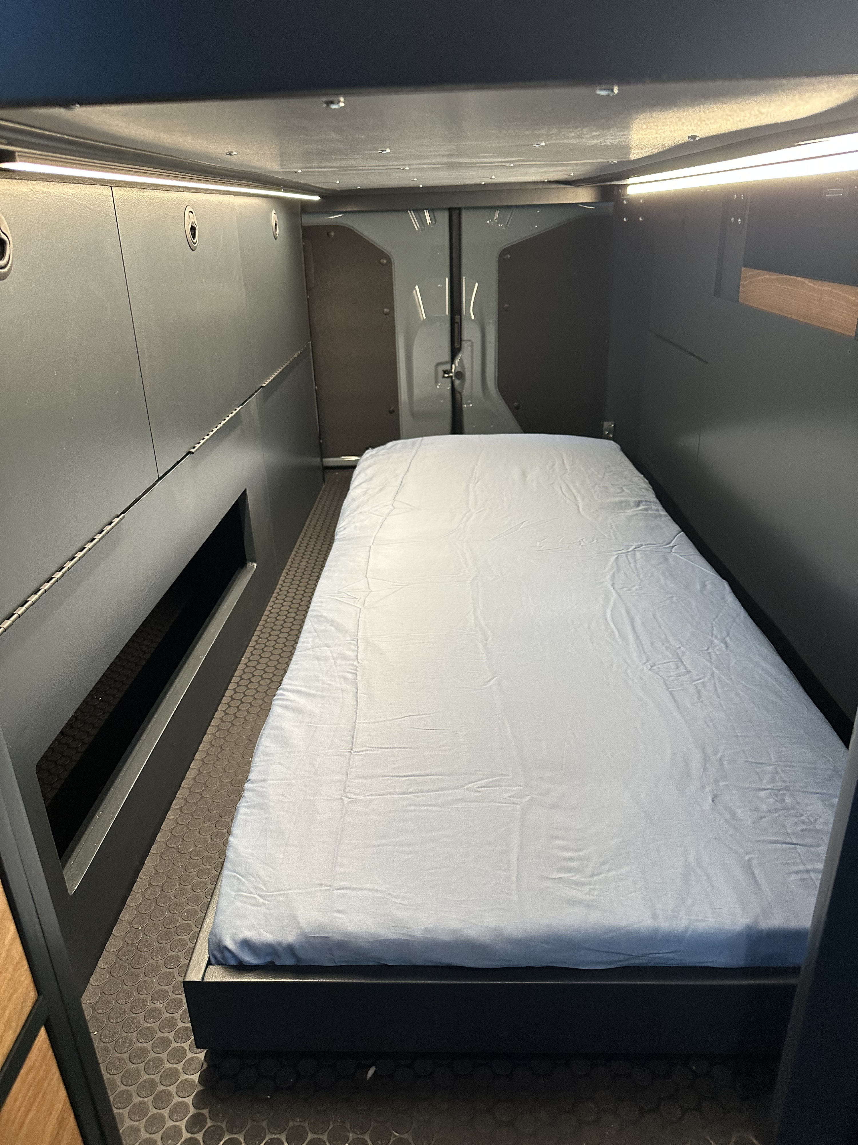 Comfortable sleeping area in a modern RV rental. Features a bed, dark walls with integrated lighting, and textured floor.. Mercedes Sprinter 2024