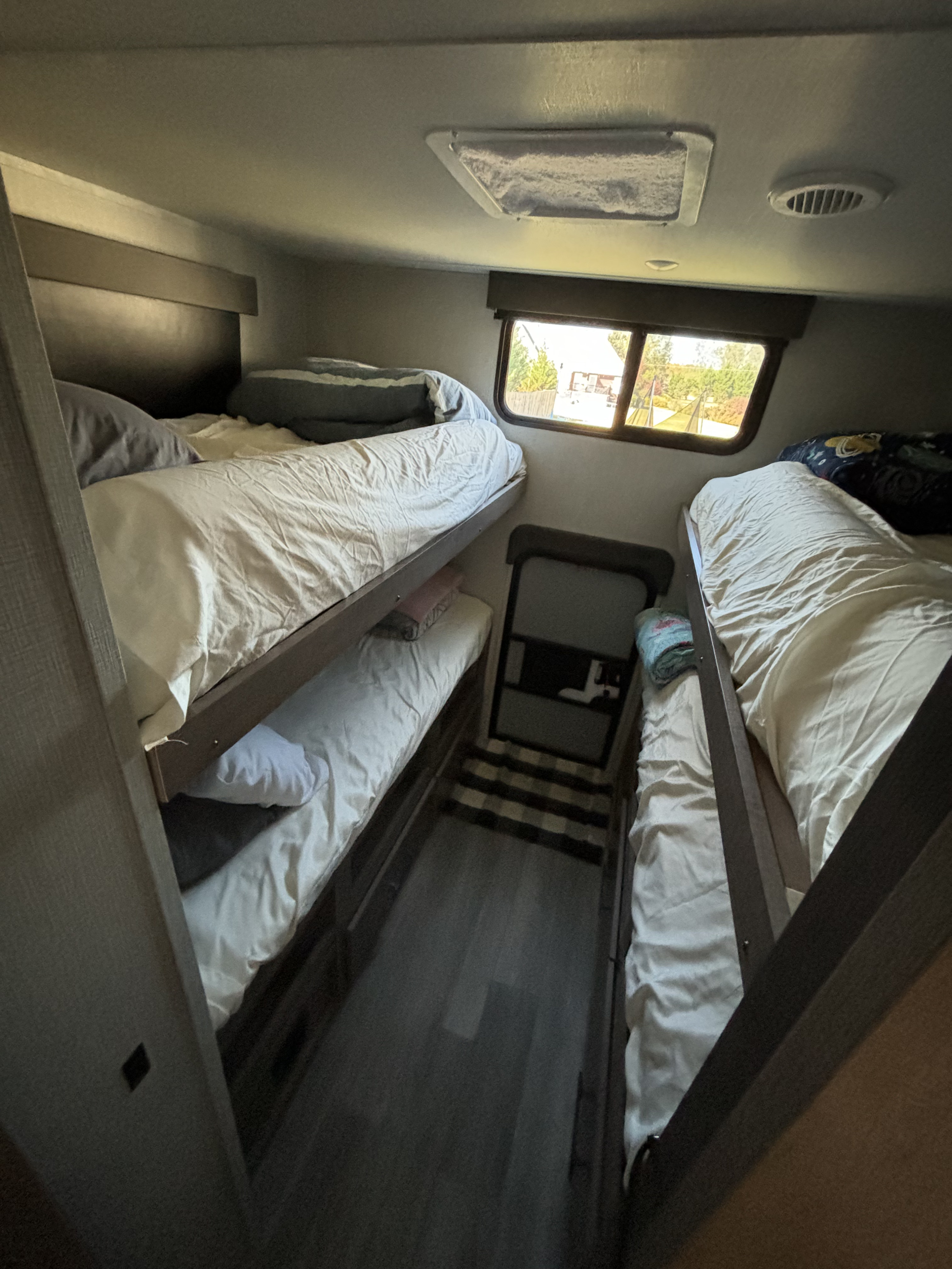 Inside an RV rental: multiple bunk beds line a narrow hall, providing a compact yet functional sleeping space with a window view.. Grand Design Imagine XLS 2023