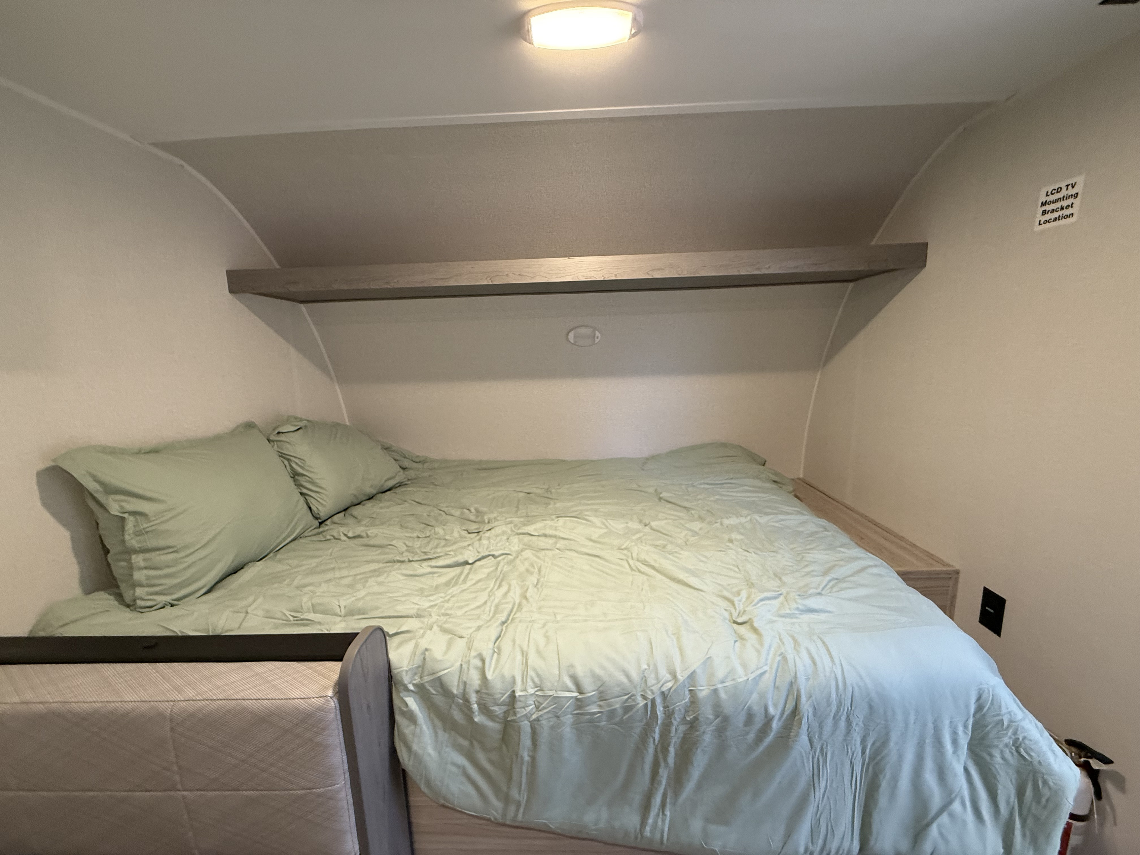 Compact rv rental bedroom with a bed, light green bedding, overhead shelf, and an LCD TV mounting label.. Heartland Trail Runner 2025
