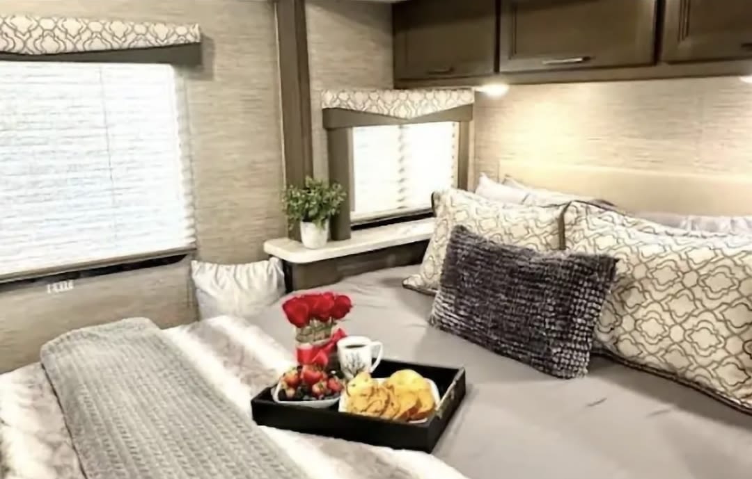 Cozy RV rental bedroom with light grey bedding, patterned pillows, & breakfast in bed on a tray.. Thor Freedom Elite 2021