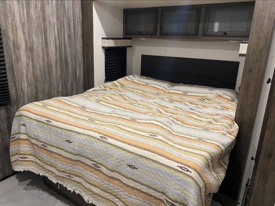 Inside an RV rental bedroom: bed with striped comforter, headboard, and dark wood overhead cabinets.. heartland Milestone 2021