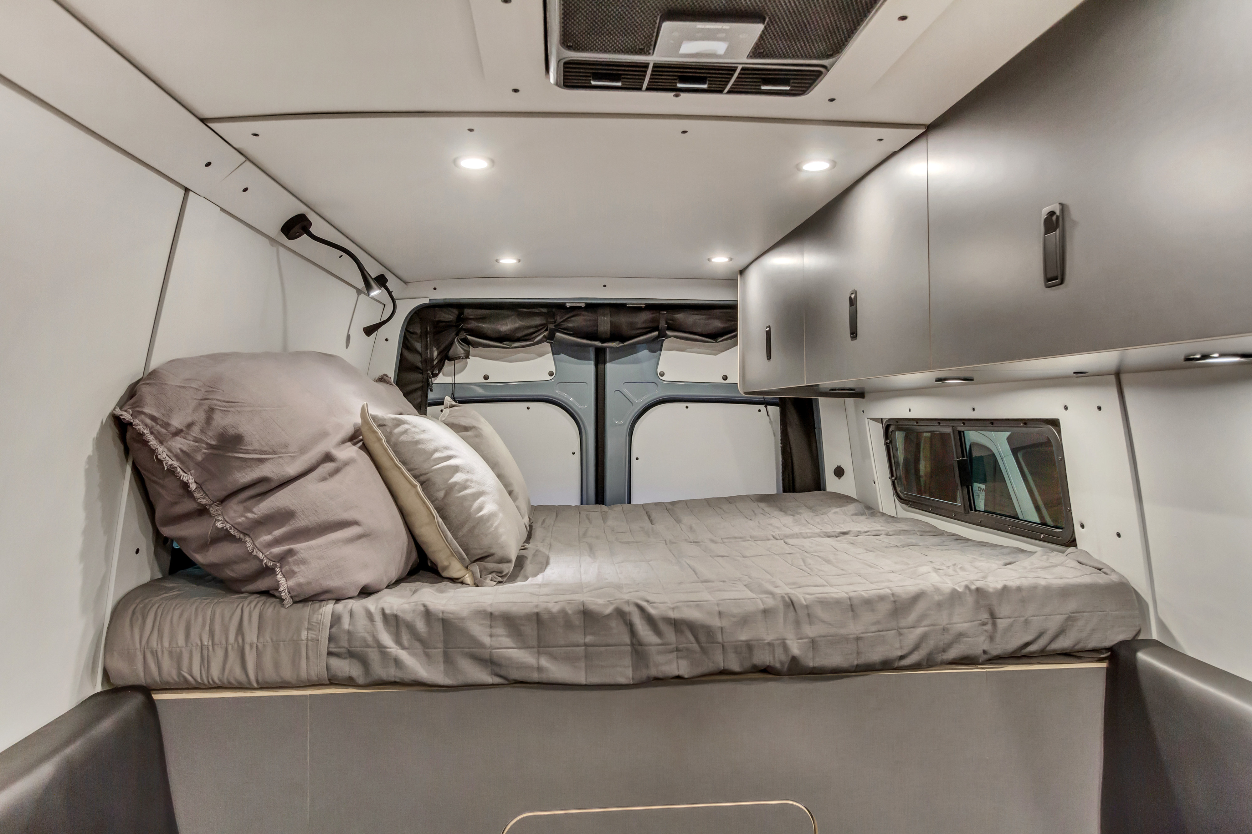 Step inside this RV rental's sleeping quarters, featuring a soft bed with pillows, sleek overhead cabinets, and bright lighting.. Mercedes-Benz Sprinter 2024