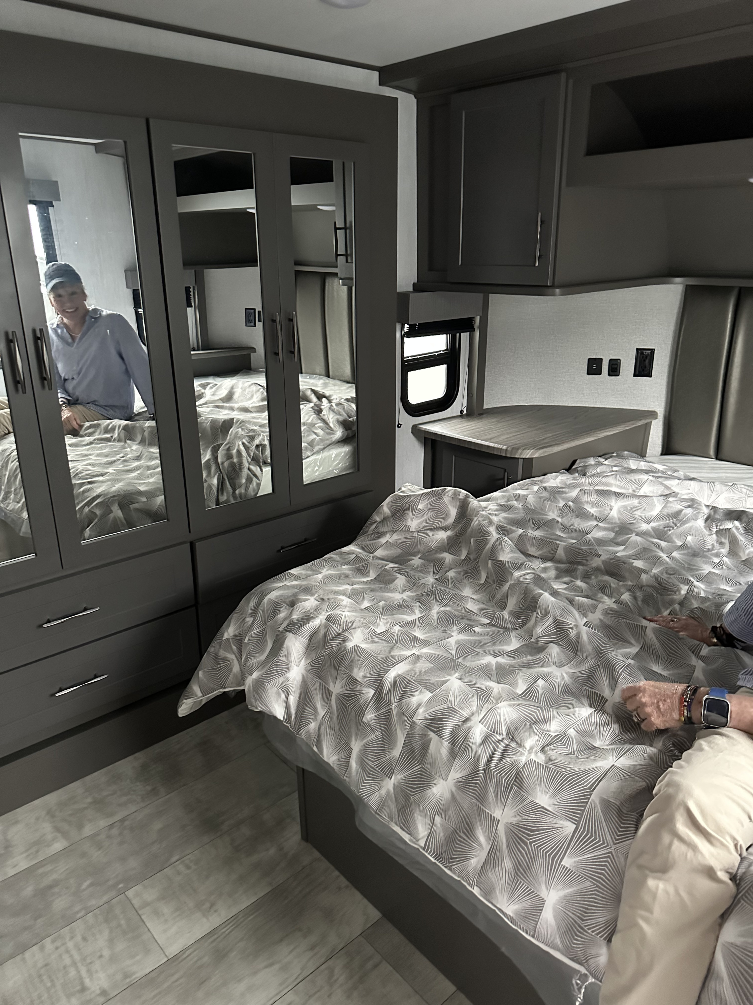 RV rental bedroom features a comfortable bed with patterned comforter, mirrored closet, and built-in storage. A person is seen in the reflection.. Grand Design Momentum G-Class Toy Hauler 2023