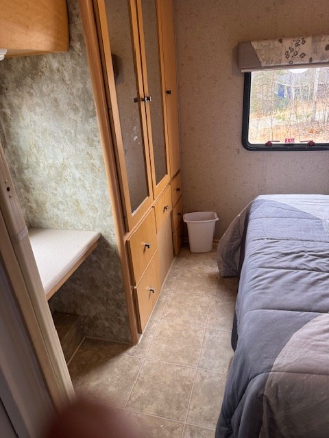 Inside an RV rental: Compact bedroom with gray bed, built-in wood cabinets, window showing trees, and tiled floor.. Winnebago Outlook 2008