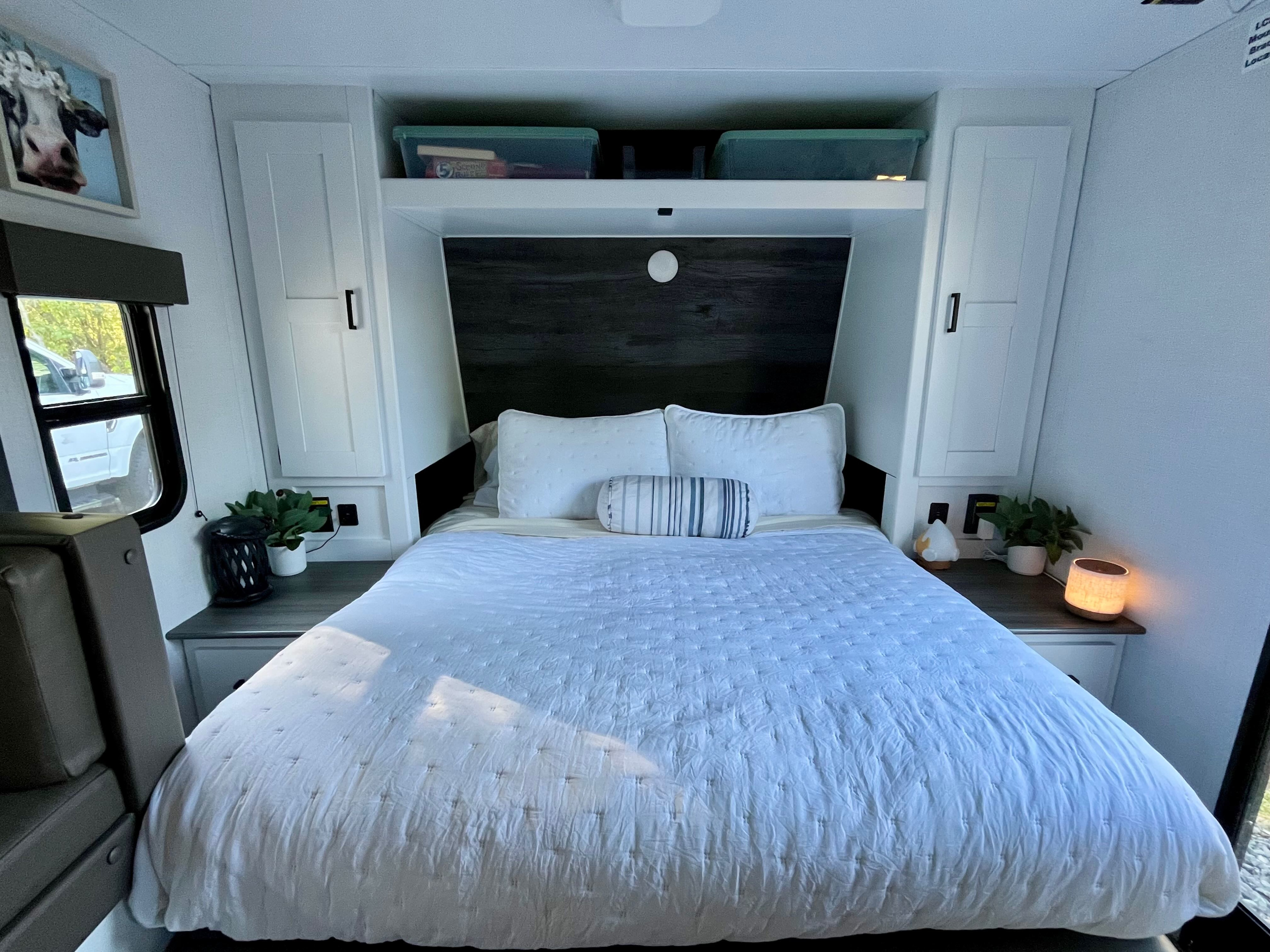 Cozy RV rental bedroom interior with a comfy bed, overhead storage, bedside lamps, and a unique cow art piece.. Keystone RV Hideout 2023