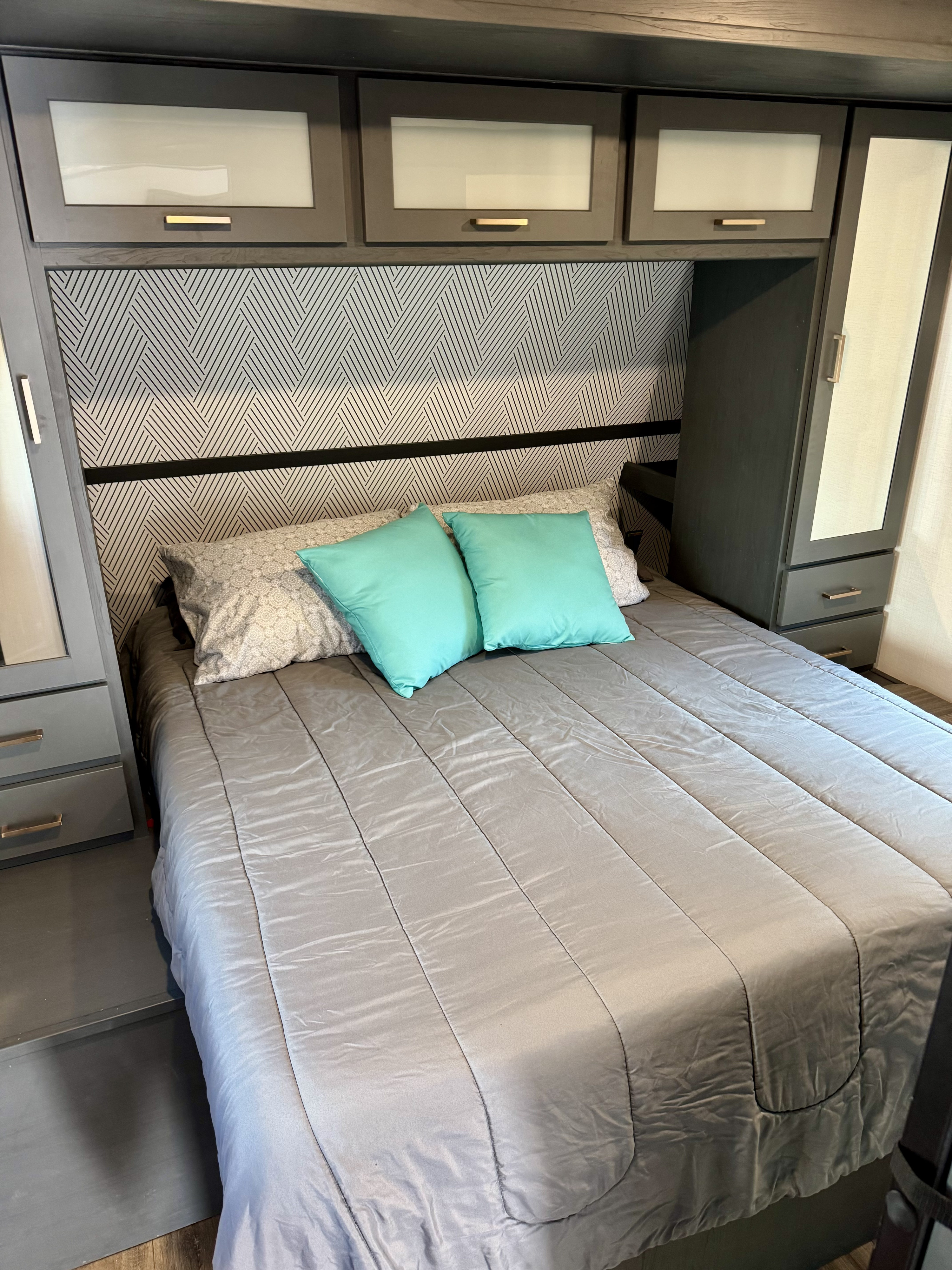 RV rental bedroom featuring a grey bed, teal pillows, geometric headboard, and sleek overhead/side storage cabinets.. Keystone RV Bullet Premier Ultra Lite 2022