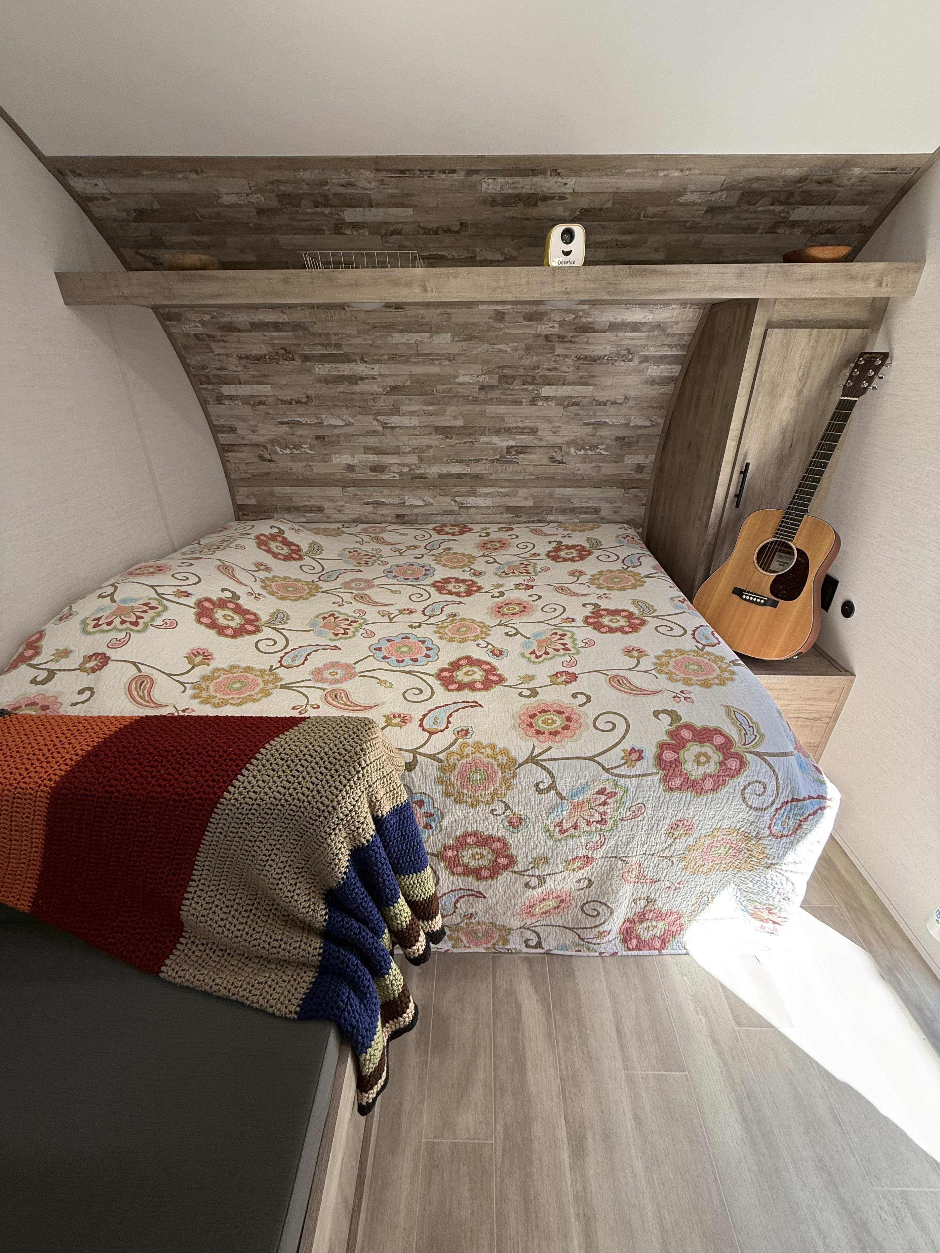 A cozy RV rental bedroom features a floral bed, knitted blanket, acoustic guitar, and an overhead shelf.. Gulf Stream Enlighten 17BH 2025