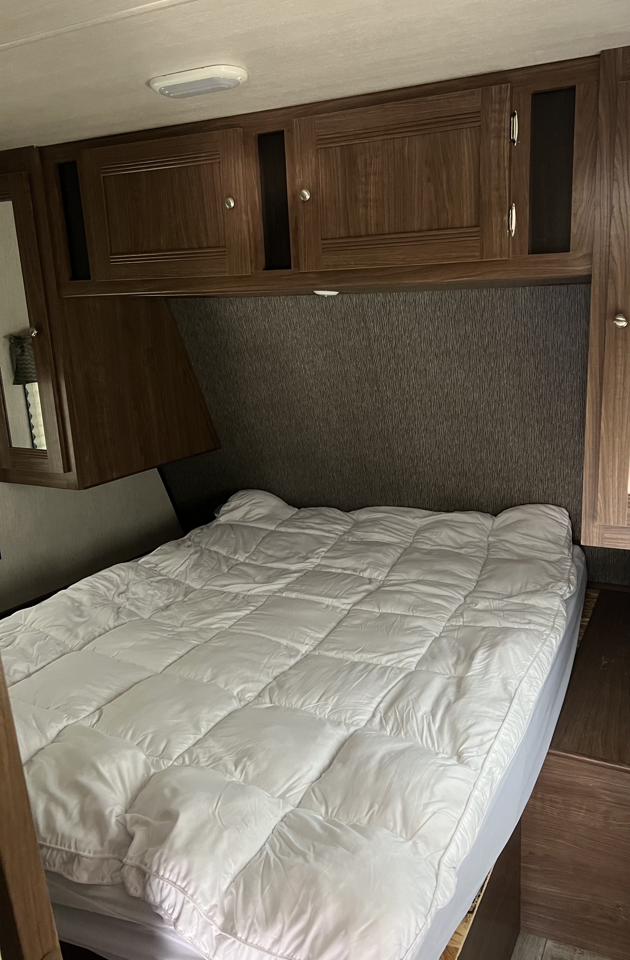 Inside an RV rental: a cozy bedroom with a white-covered bed, dark headboard, and wooden cabinets overhead. Ready for your trip!. Heartland RVs Pioneer 2018