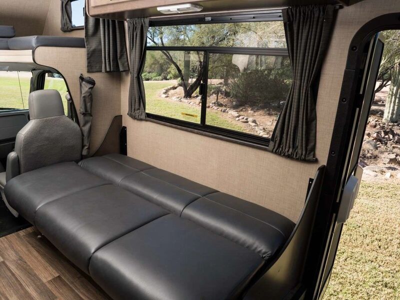RV rental interior featuring a black sofa bed, swiveled driver's seat, overhead bunk, and a scenic window view.. Thor Majestic 2019