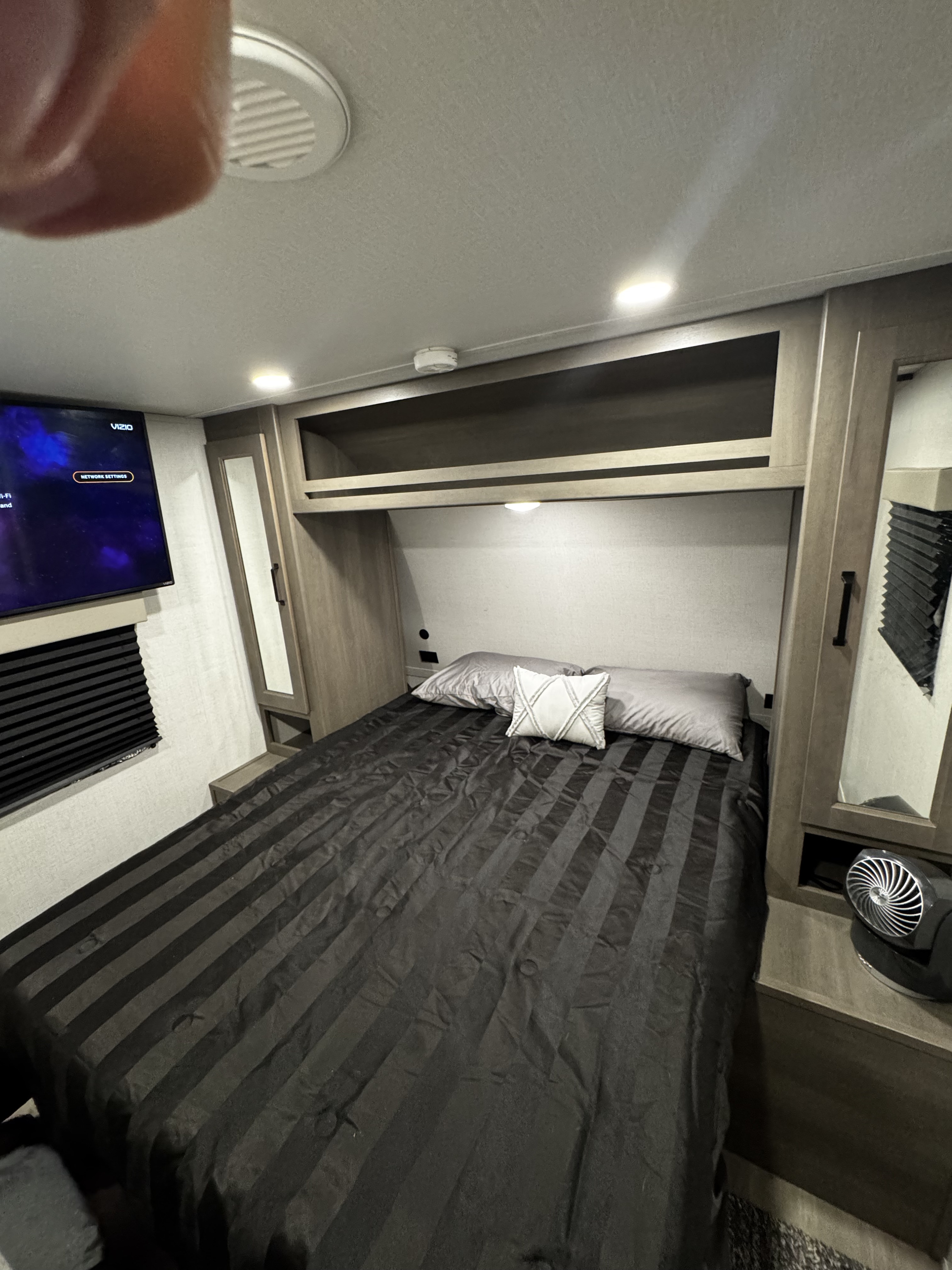 Compact RV rental bedroom showing dark striped bed, wall-mounted TV, and side storage with a small fan.. Grand Design Transcend Xplor 2024