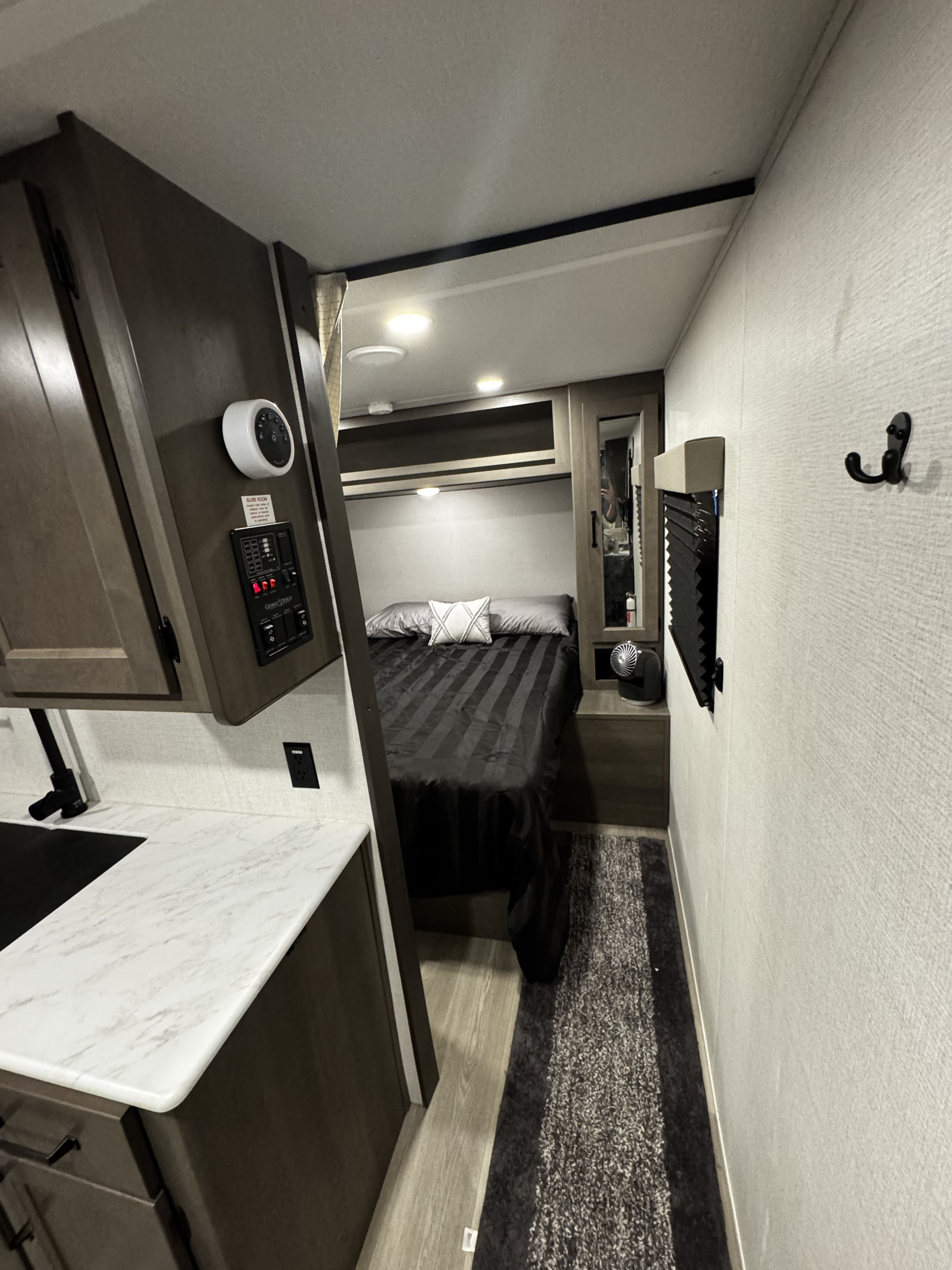 View of an RV rental bedroom with a striped bedspread, overhead cabinets, and adjacent kitchen counter with control panel.. Grand Design Transcend Xplor 2024