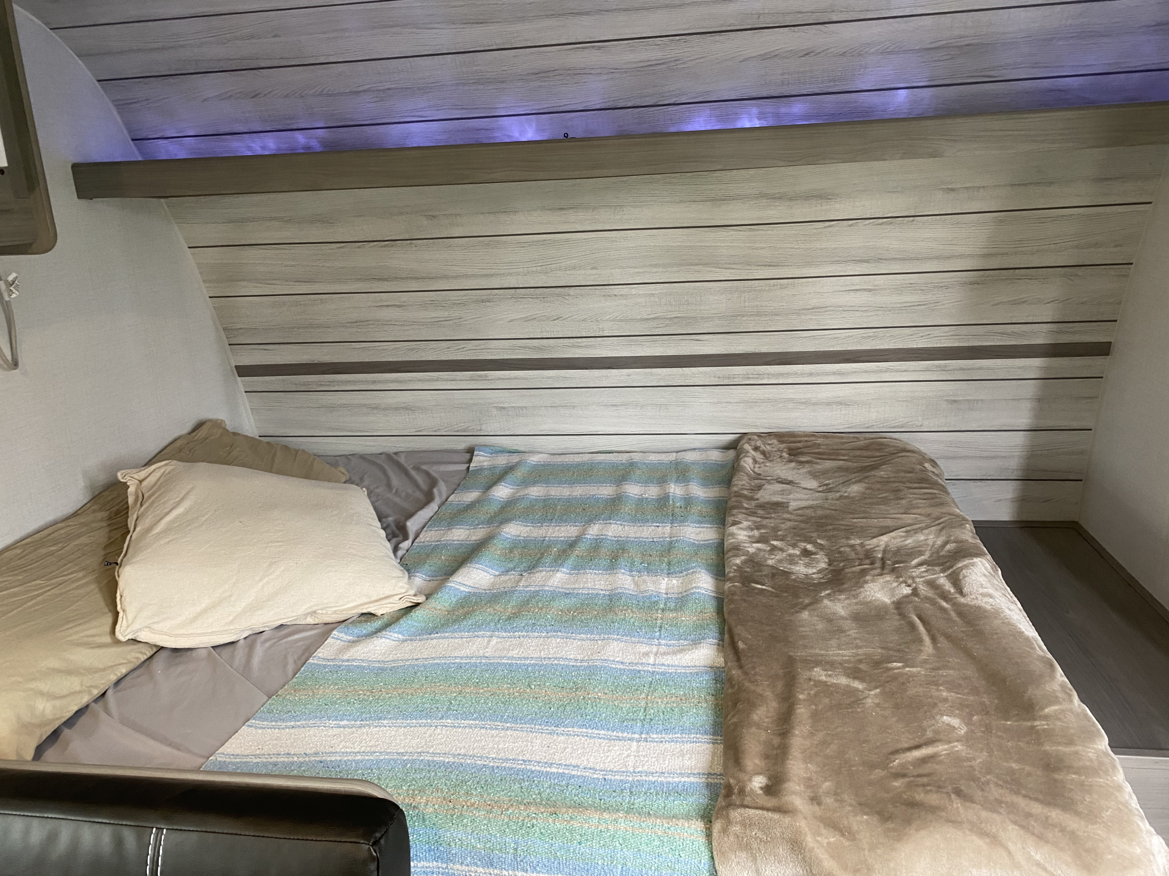 Cozy RV rental bed with wood-grain paneling, subtle purple LED lighting, pillows, and colorful blankets for comfort.. Coleman Lantern 17B 2024