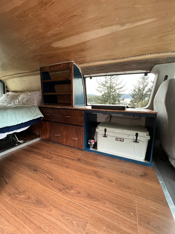 Inside an RV rental van conversion: Custom storage, bed, wood floors, and a Yeti cooler. Scenic window view.. Ford Ford E350 2006