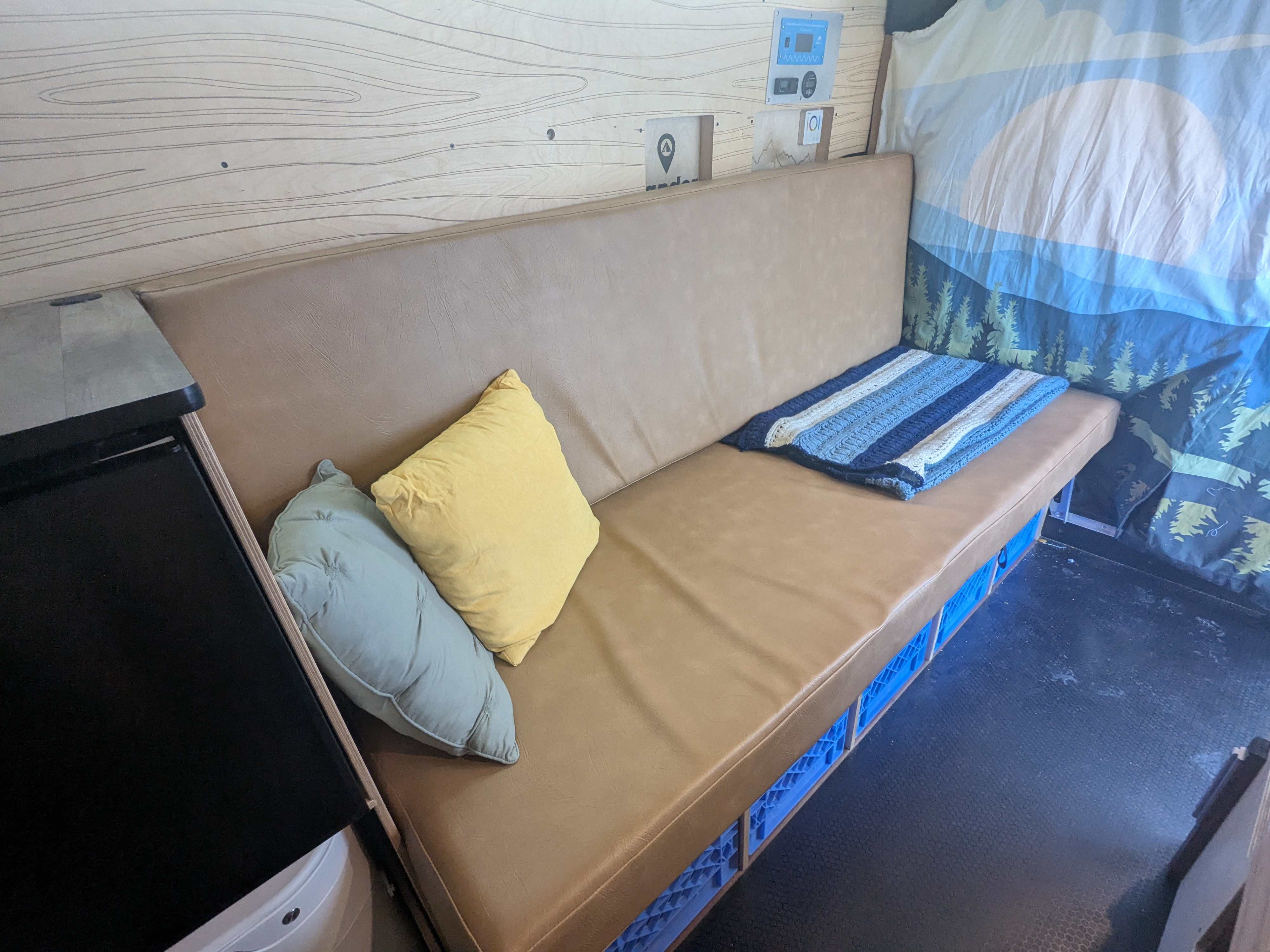 RV rental interior: Light brown bench, pillows, striped blanket, blue crate storage, wood wall, and control panel.. RAM Promaster 2023