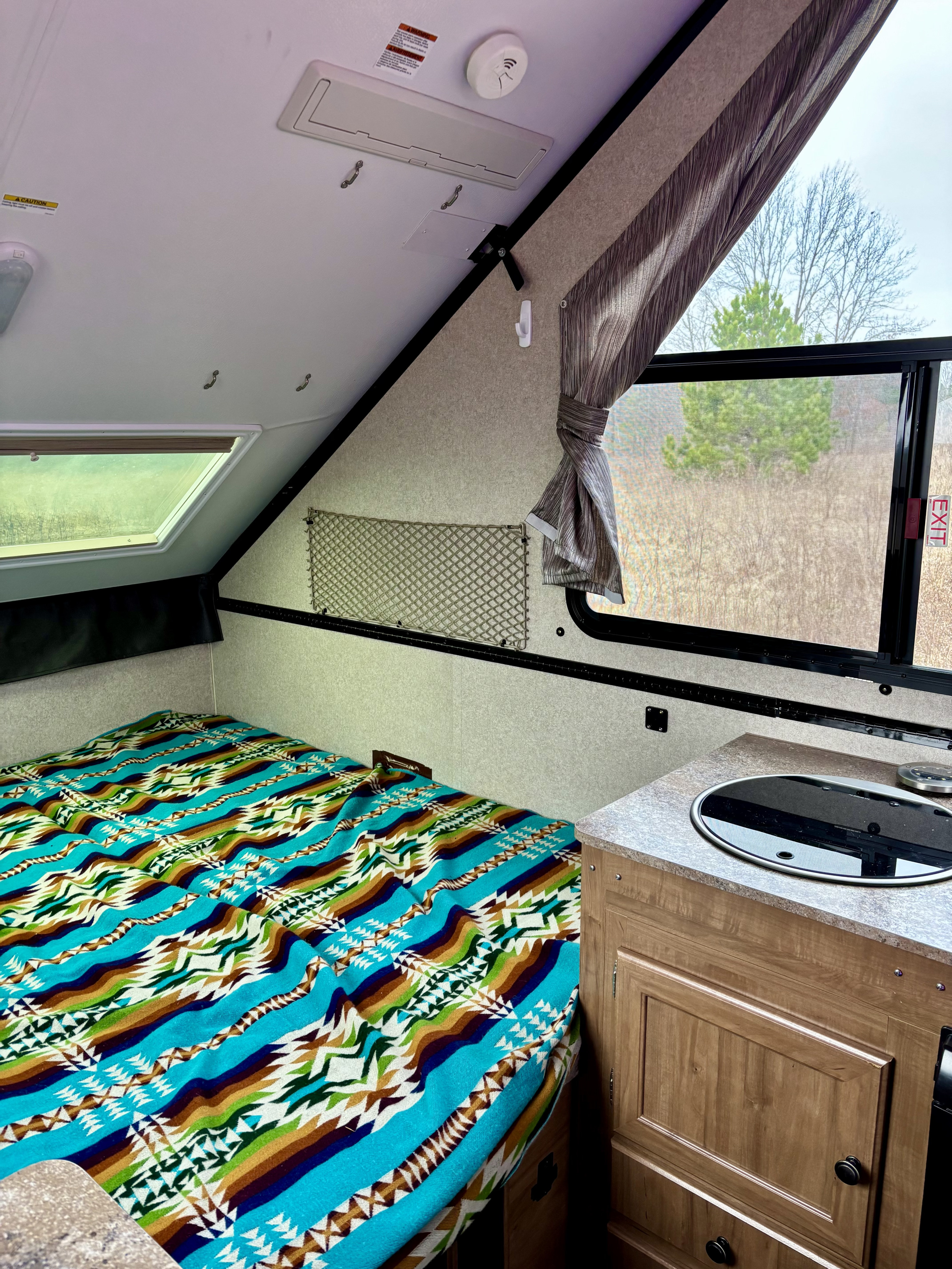 Cozy RV rental interior with bed, patterned blanket, compact kitchen, and window view for comfortable travel.. Forest River Rockwood Premier 2019
