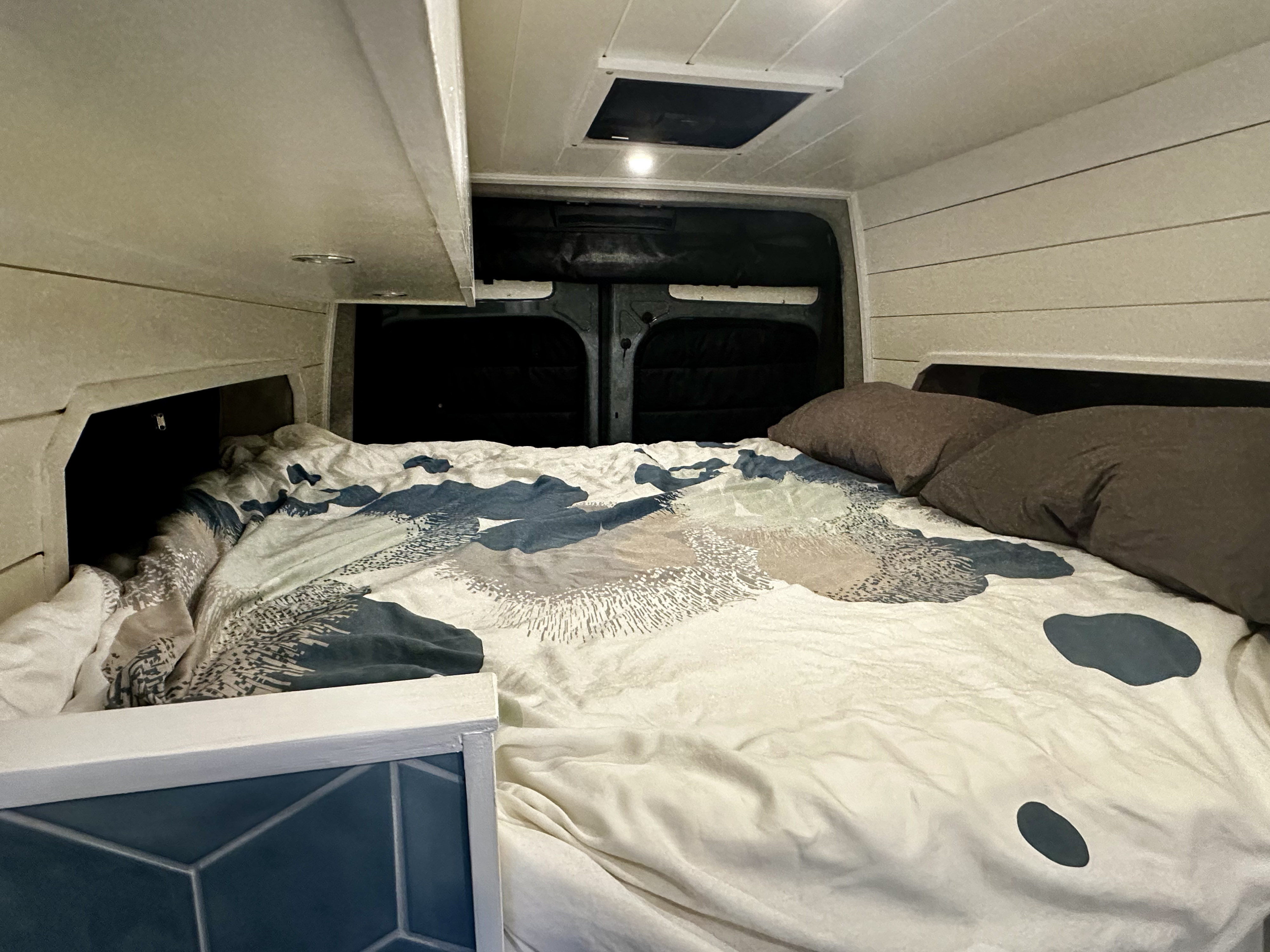 An RV rental's cozy sleeping area features a bed with patterned bedding, built-in storage, and a ceiling vent.. Mercedes-Benz Sprinter 2021