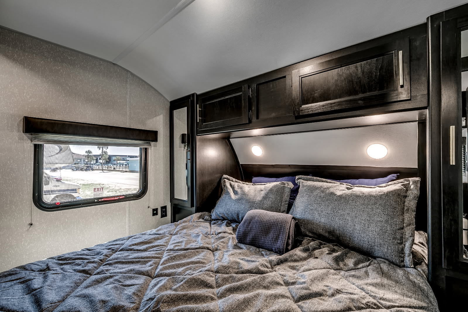 Inside a modern RV rental bedroom: a cozy bed with gray bedding, dark overhead storage, and a window with an "EXIT" sign and outdoor view.. Cherokee Grey Wolf 2022