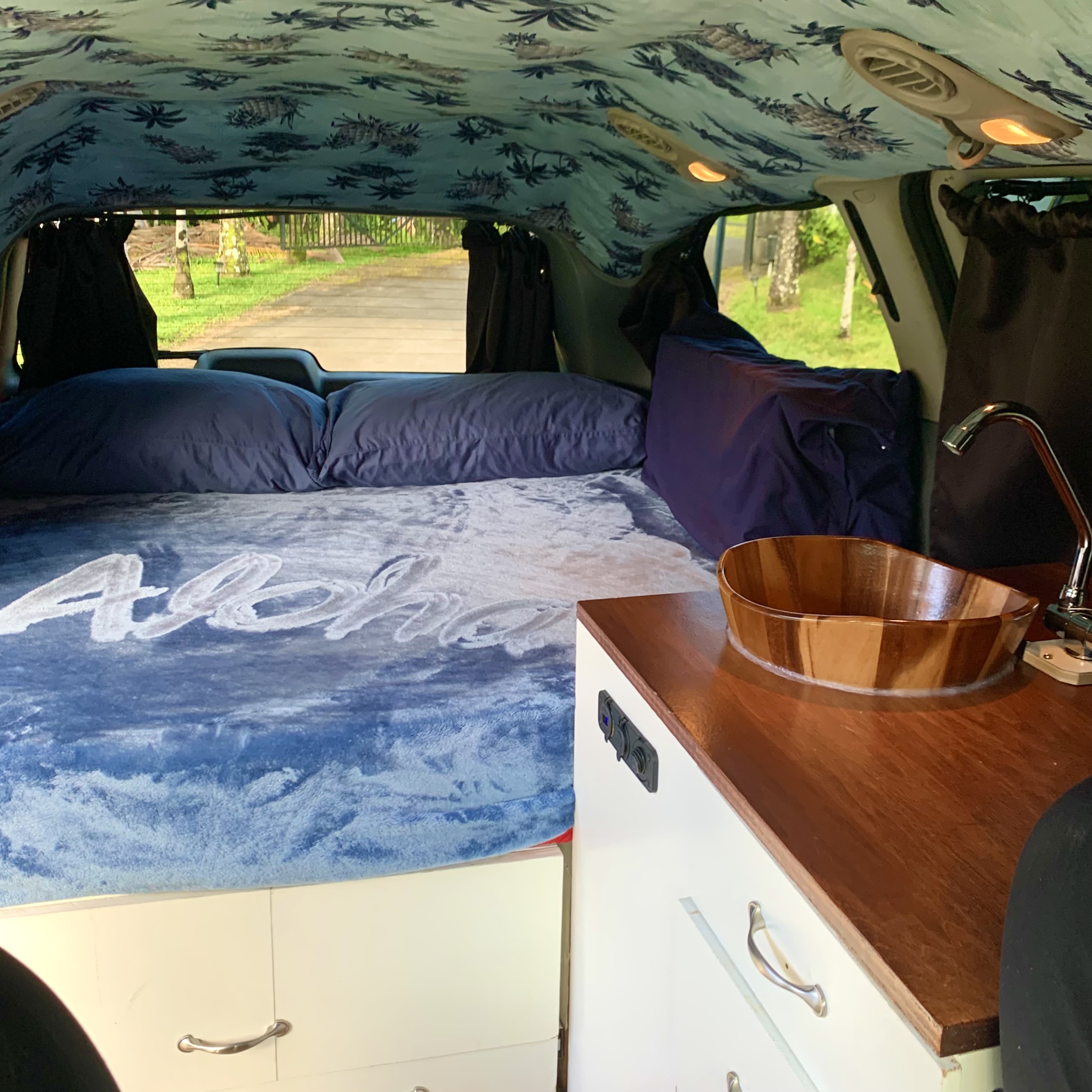 Cozy RV rental interior: bed with "Aloha" blanket, custom ceiling, and small sink for comfortable mobile living.. Dodge Grand Caravan 2007