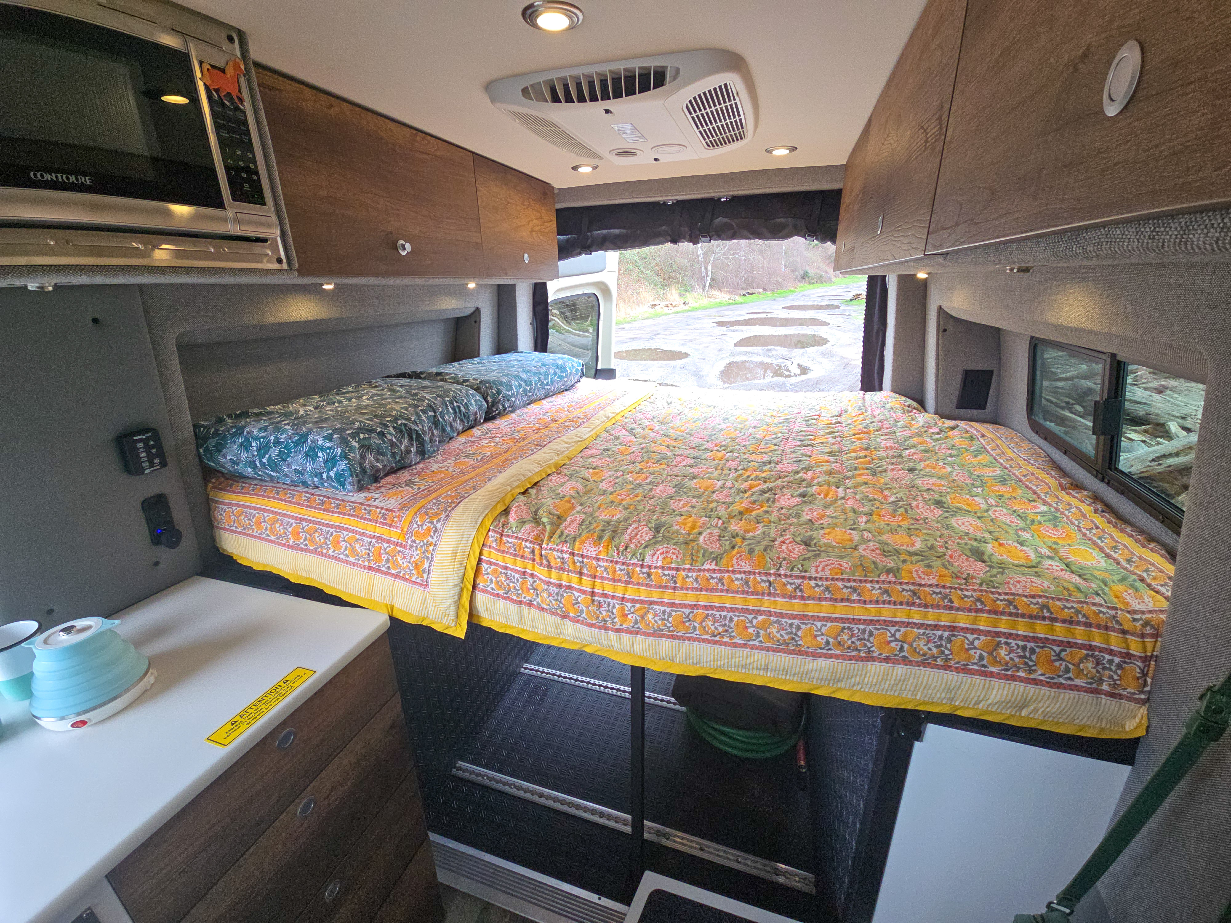 Cozy RV rental interior with a bright bed, microwave, and kettle. Open rear view reveals a wet road.. Mercedes Sprinter 2021