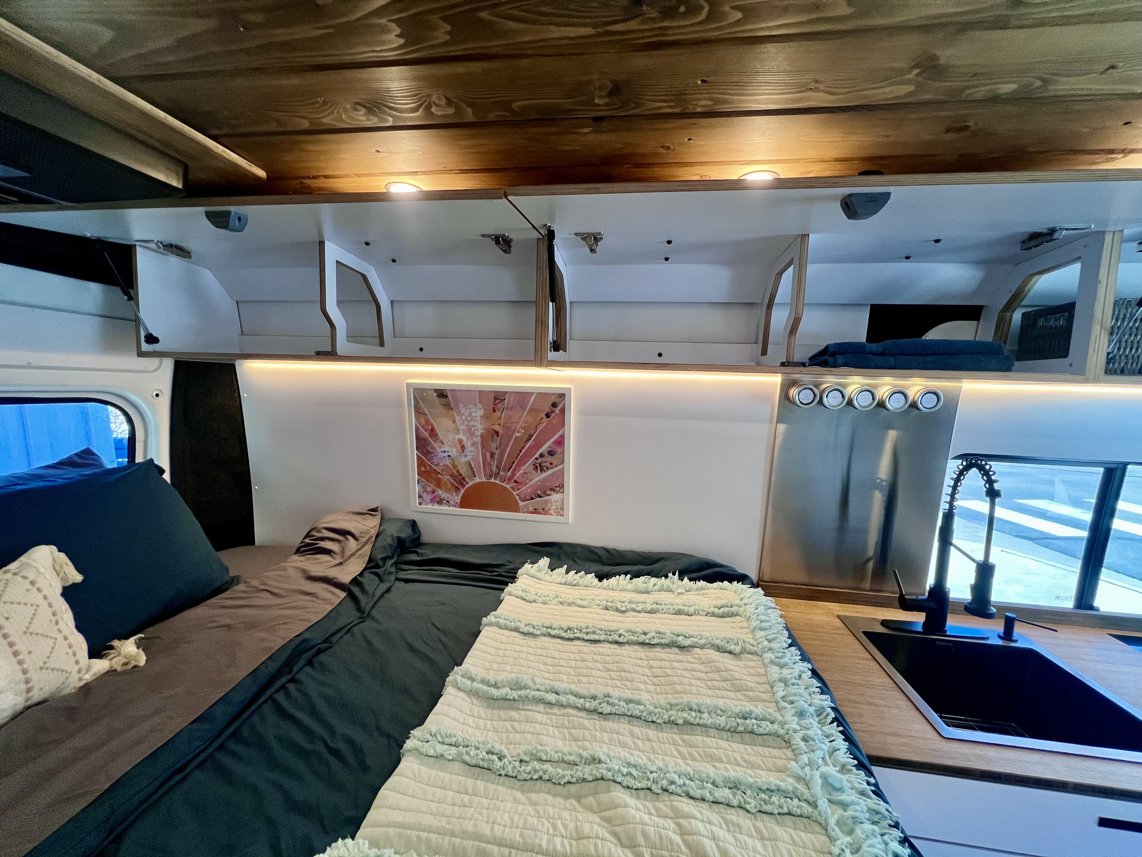 An rv rental's cozy interior: bed with pillows, open overhead storage, warm LED lighting, and a compact kitchen.. Dodge Ram Promaster 2500 159 2020