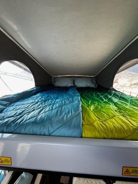 Inside an RV rental, a comfortable pop-top bed with vibrant blue and green sleeping bags, pillows, and bright windows.. Winnebago Solis 2023
