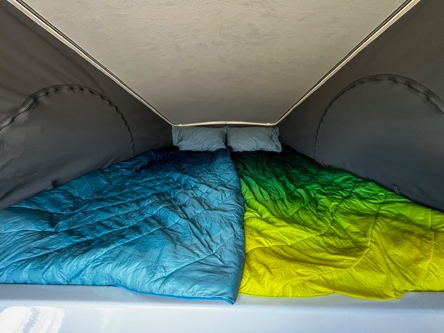 Inside a pop-top RV rental: a cozy bed with bright blue & green-yellow comforters and pillows. Ready for adventure.. Winnebago Solis 2023
