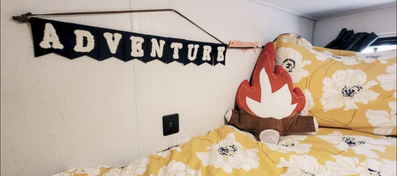 Inside an RV rental, a comfy bed with yellow floral bedding, an "ADVENTURE" banner, and a campfire pillow create a cozy space.. Forest River Wildwood FSX 2022