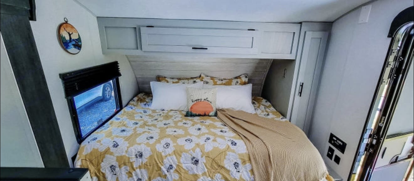 Step into comfort! This cozy RV rental bedroom features cheerful floral bedding, overhead storage, and a scenic window view.. Forest River Wildwood FSX 2022