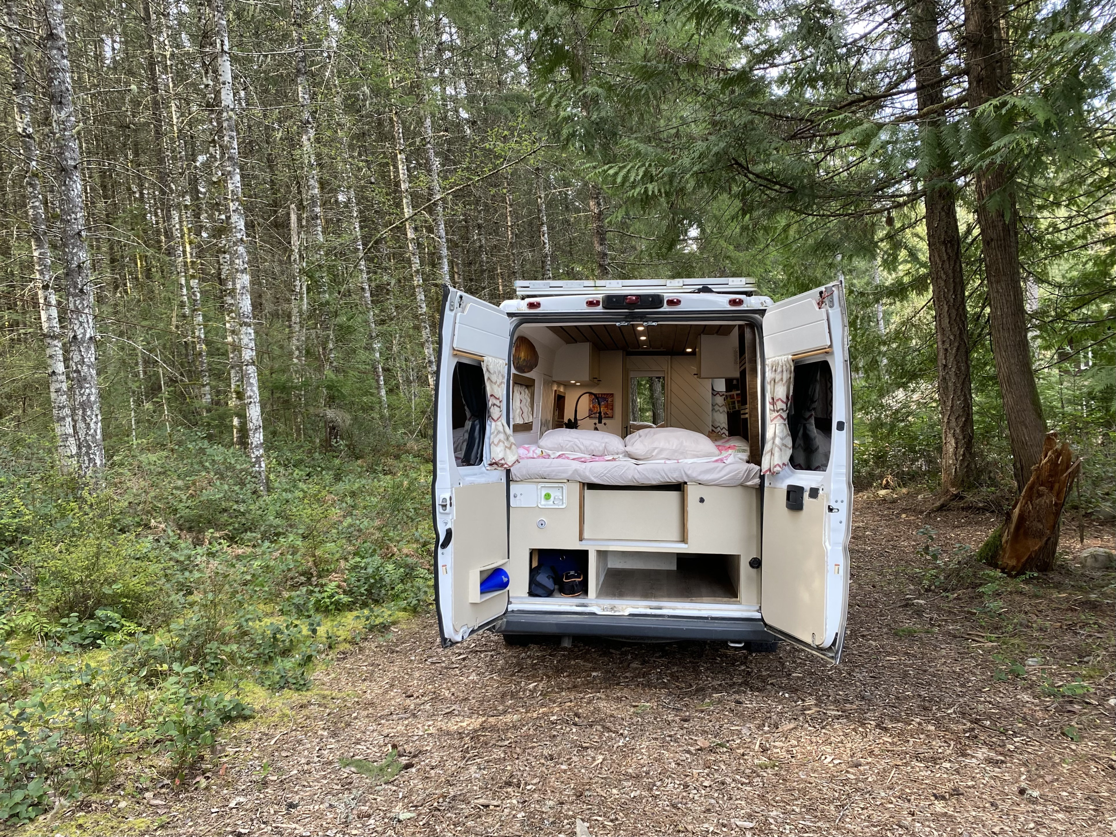 Open RV rental rear doors reveal a cozy bed & kitchen, parked on a forest path. Perfect for van life camping adventures.. Ram Promaster 2018