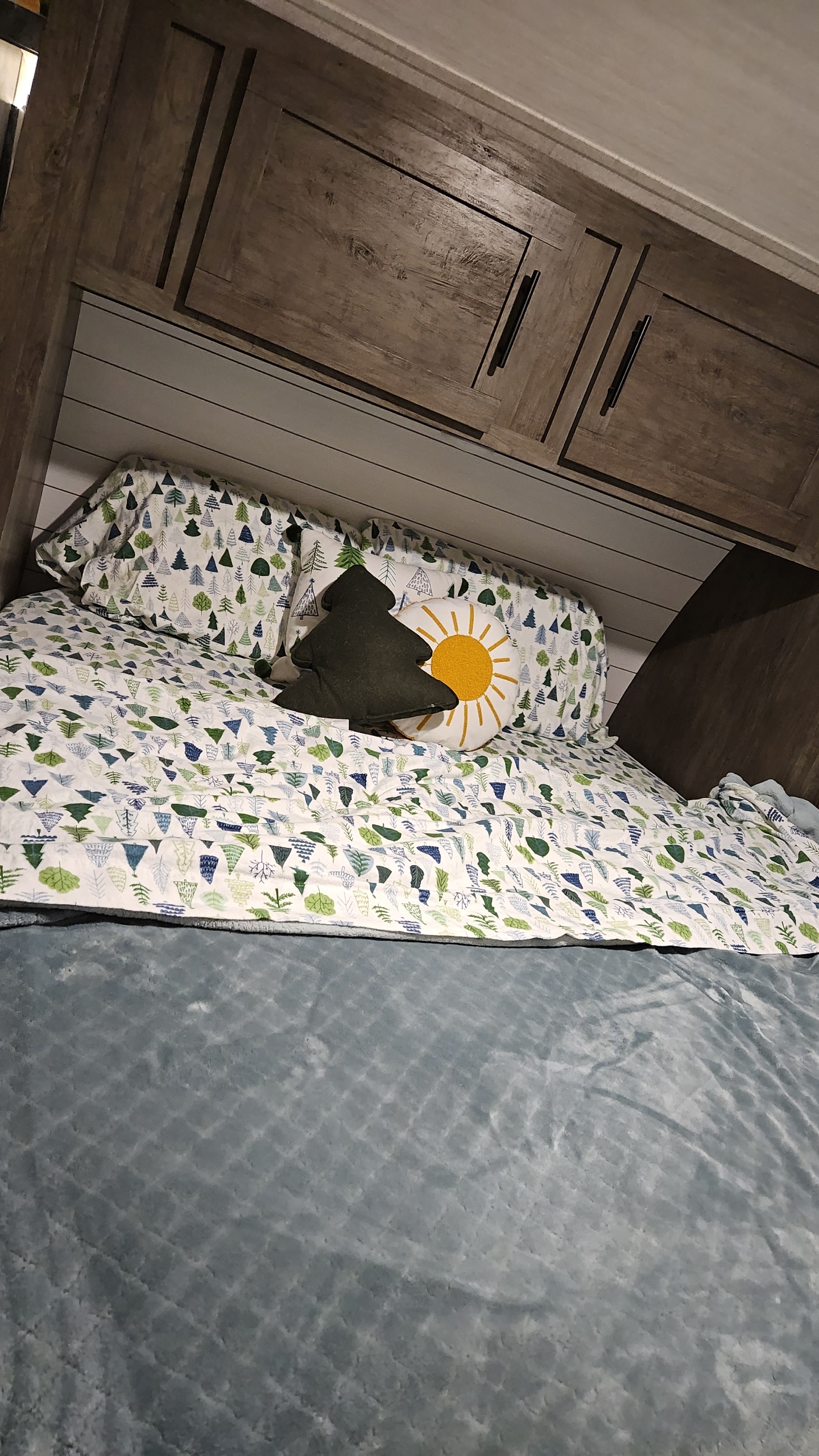Cozy bed in an RV rental with tree-patterned bedding, a tree pillow, and a sun pillow, under wood cabinets & shiplap.. Forest River Salem 2021
