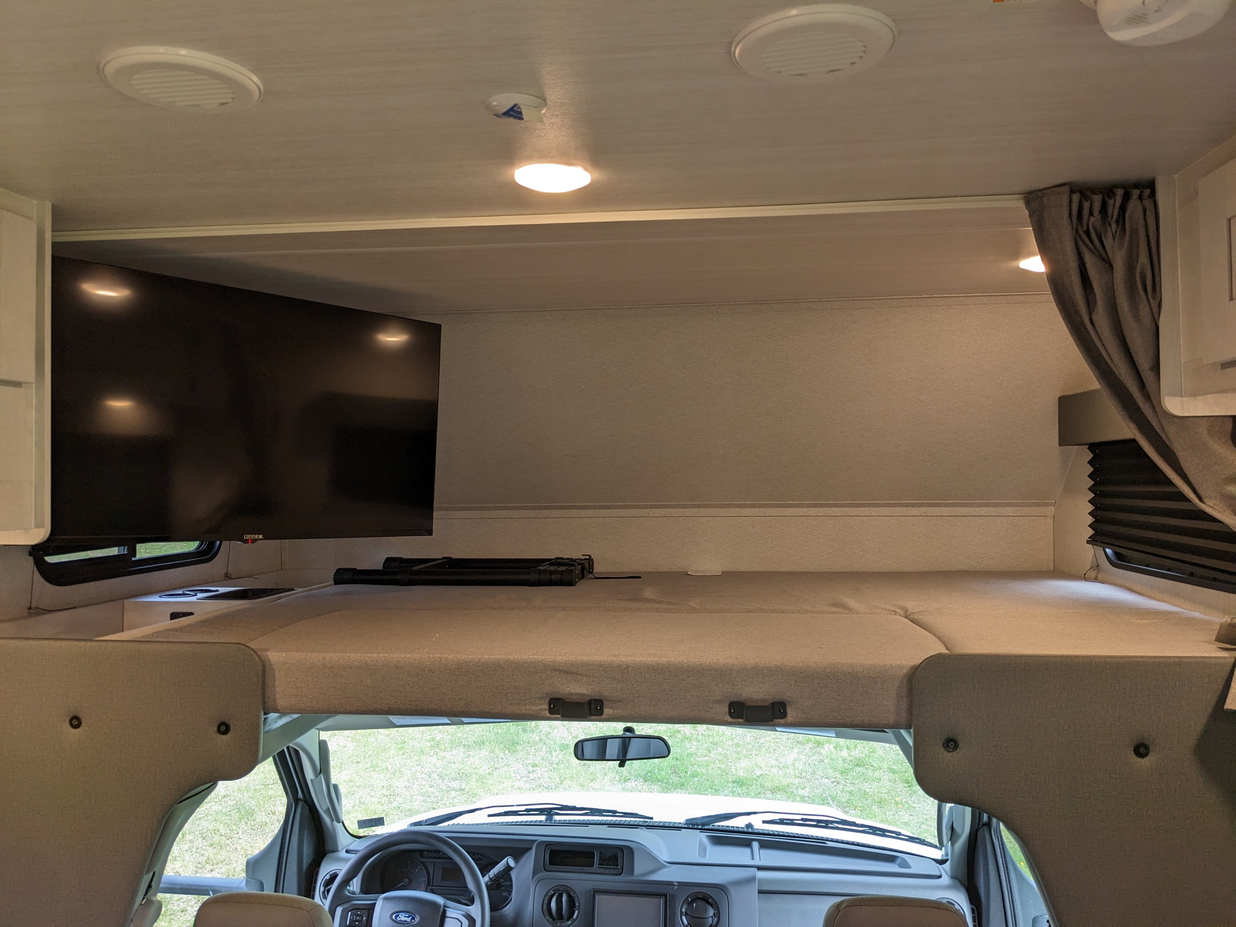 Interior view of an RV rental: cozy overhead bunk with TV, looking into the driver's cab.. East to West Entrada 2026