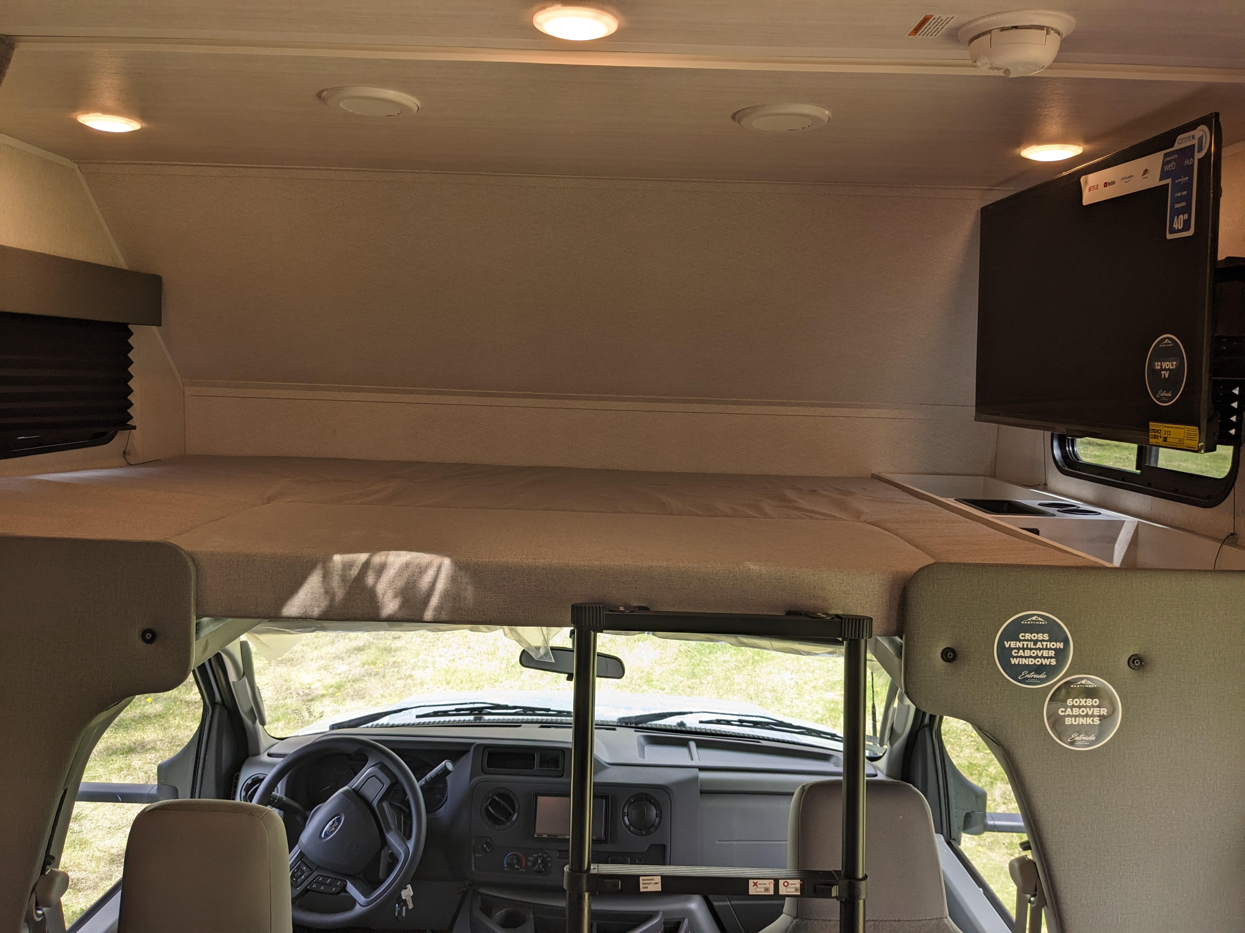 Inside an RV rental, featuring a cozy cabover bunk bed, a mounted TV, and the vehicle&#x27;s dashboard below.. East to West Entrada 2026