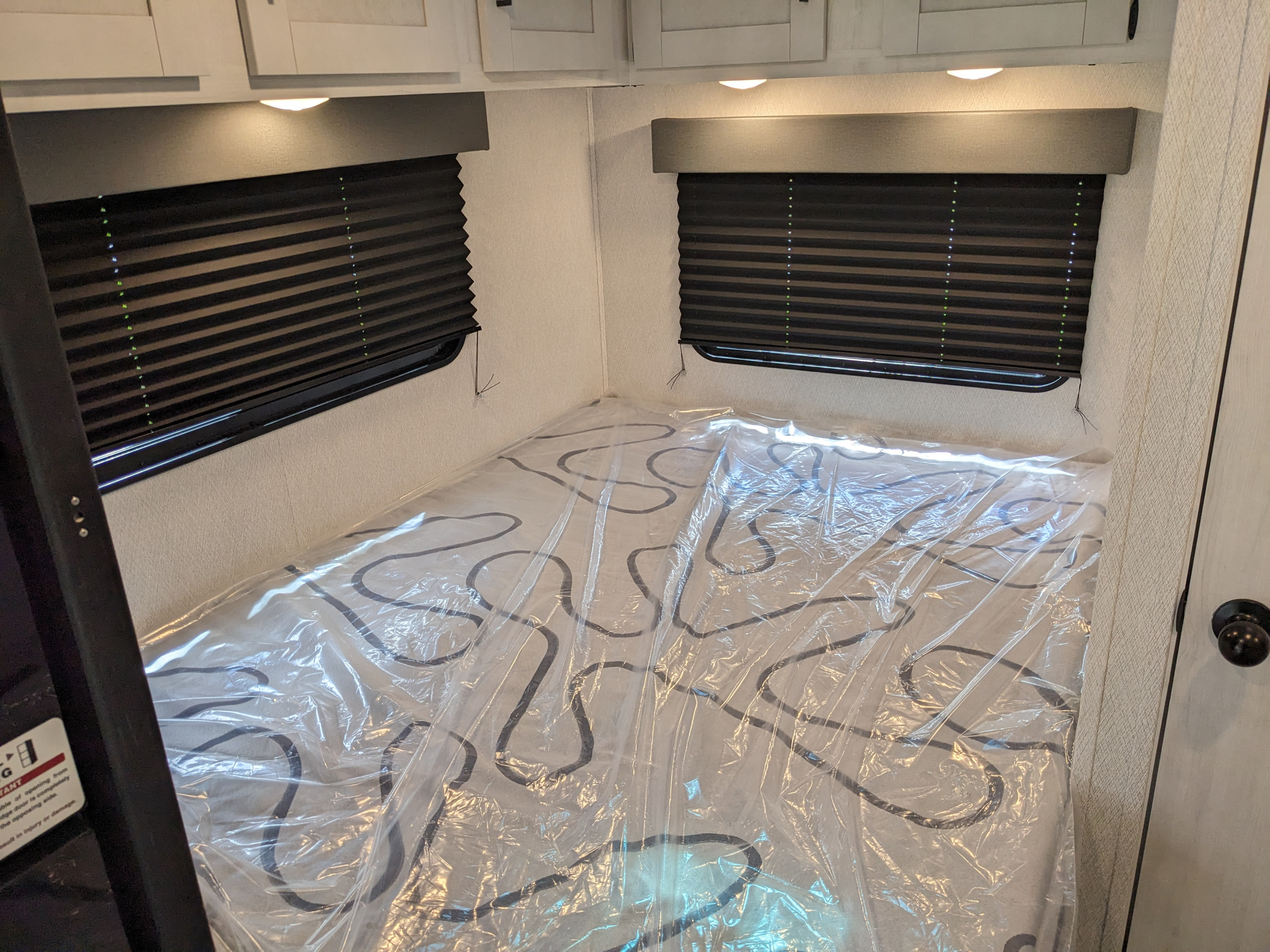 RV rental bedroom shows a new, plastic-covered patterned mattress, dark window blinds, and light overhead cabinets.. East to West Entrada 2026