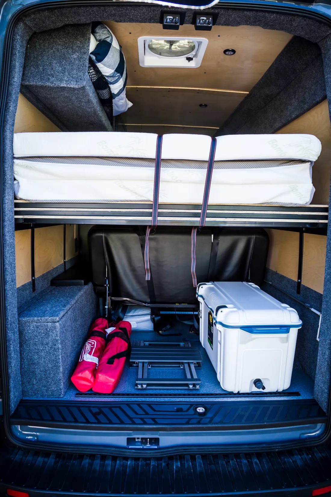 Rear view of an RV rental: features a high bed platform with stacked mattresses, a seat, cooler, & gear.. Ford Transit 2022
