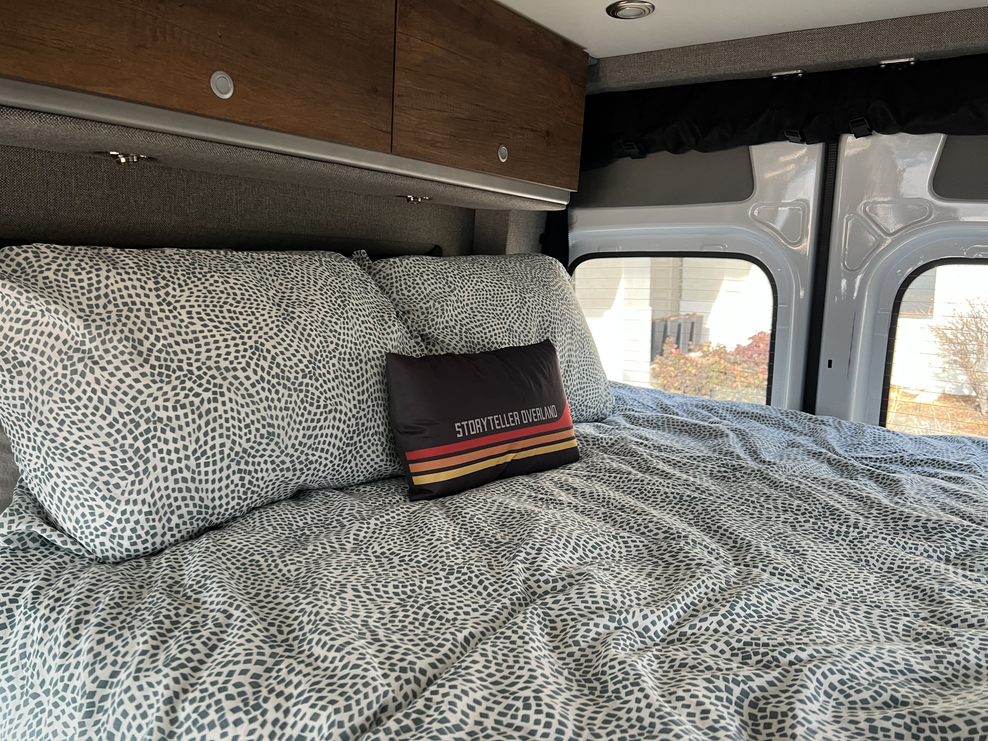 Cozy RV rental bedroom with patterned bedding, overhead cabinets, and rear windows.. Storyteller Overland Sprinter 2022