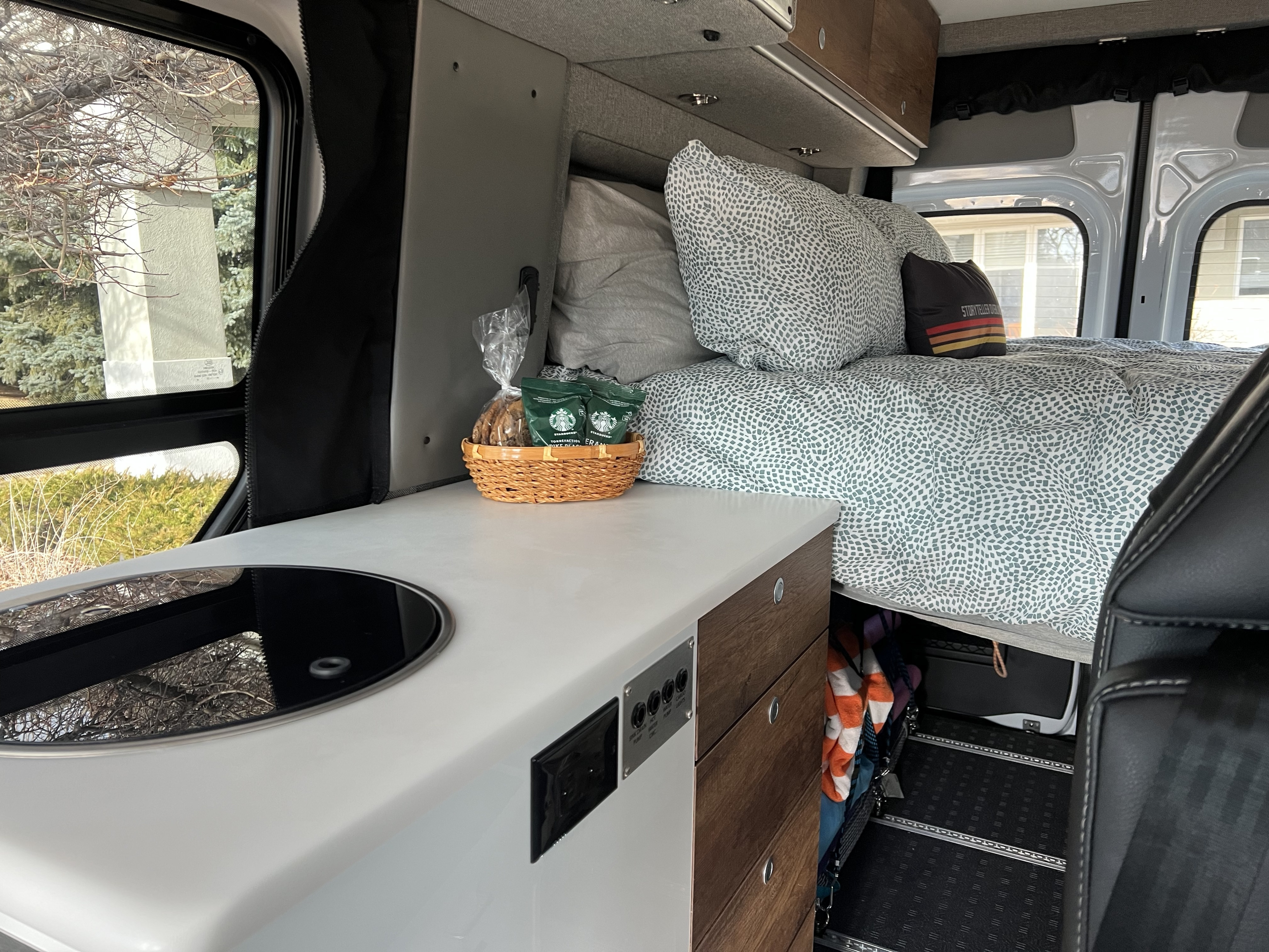 RV rental interior: comfy bed with patterned duvet, compact kitchen with cooktop, and wooden storage.. Storyteller Overland Sprinter 2022