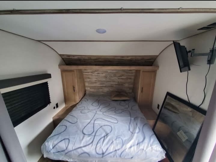 Compact & cozy RV rental bedroom with a wood headboard, comfy bed, mounted TV, and window for travel.. Gulf Stream Grand River BH25 2024