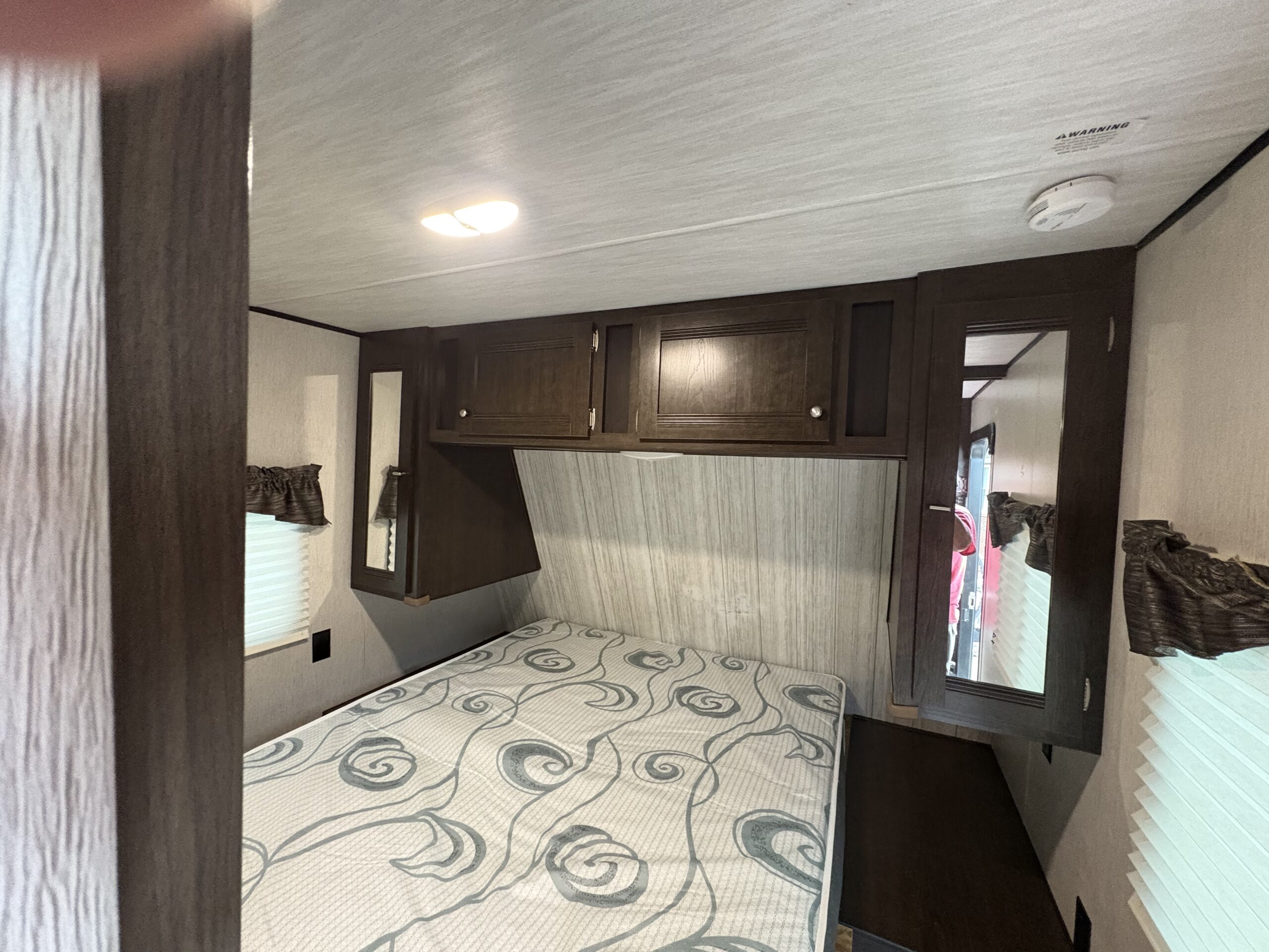 Compact RV rental bedroom with patterned mattress, dark wood cabinets, built-in mirrors, and a window.. Heartland Pioneer 2020