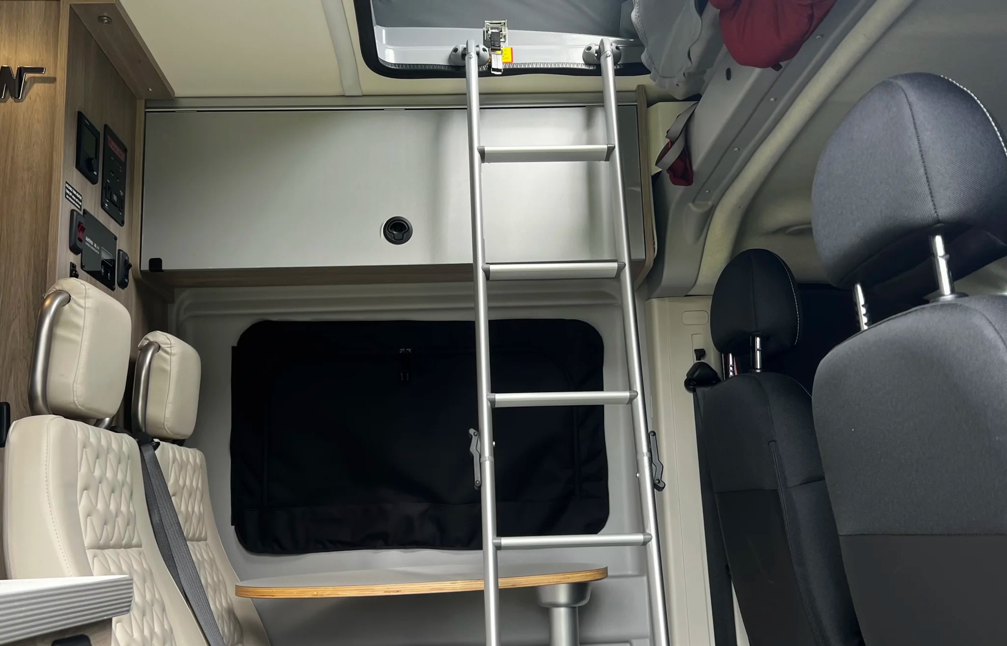 Inside this RV rental, a ladder leads to an overhead bunk. Features include comfortable seating, a small table, and control panels.. Winnebago Solis 2023
