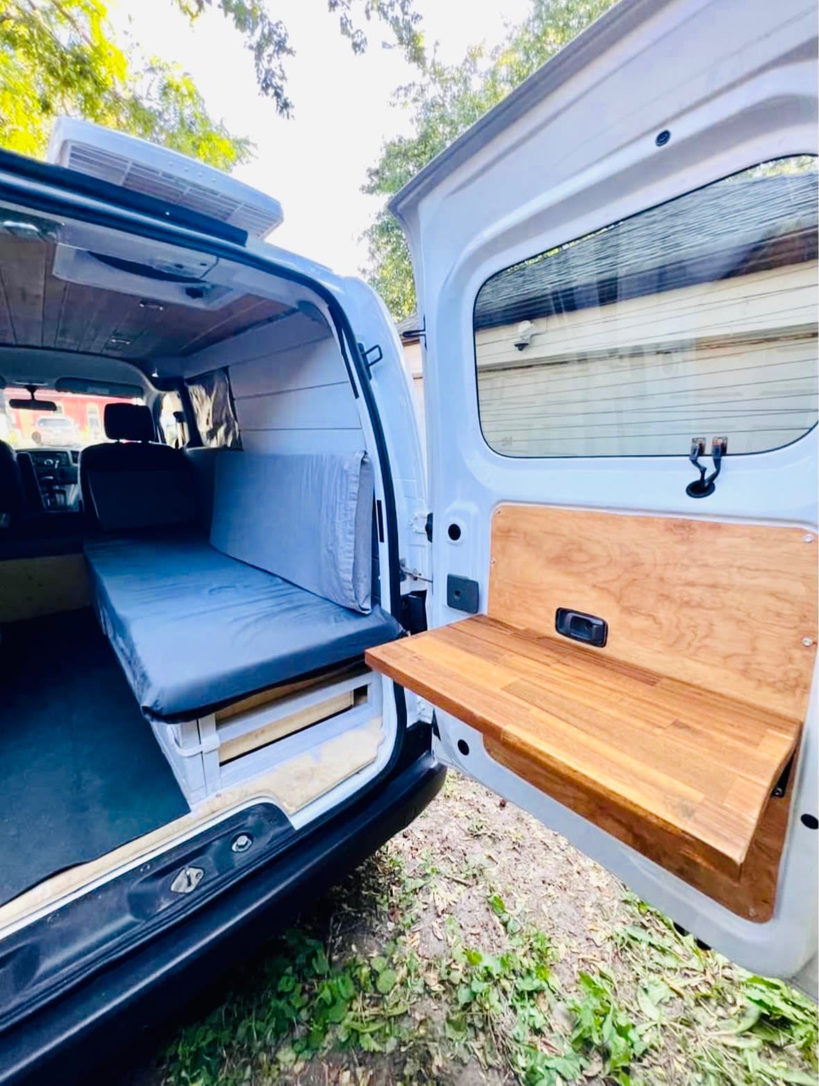 View into an RV rental camper van from the open rear doors, revealing a grey bed/bench and a practical wooden fold-out table.. Nissan NV200 2015